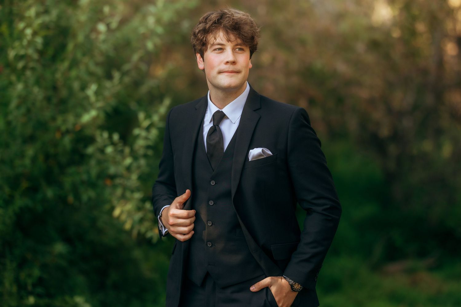 Groomsman in formal black tuxedo poses against natural foliage background.