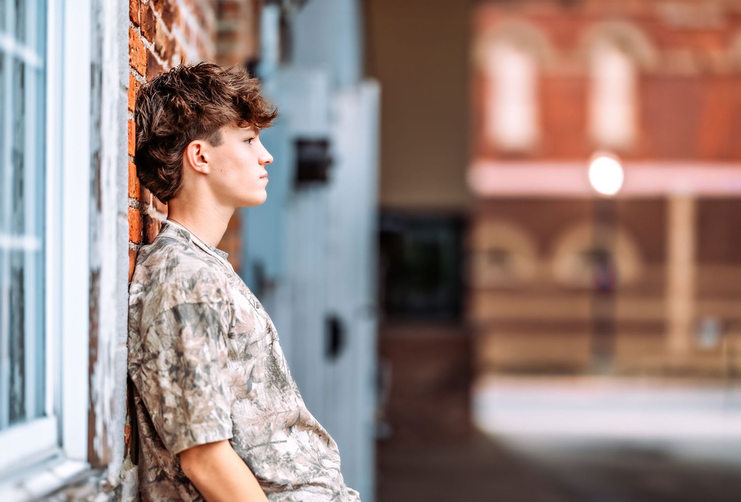 A person in a floral shirt leans against a white wall with a window in a casual pose.