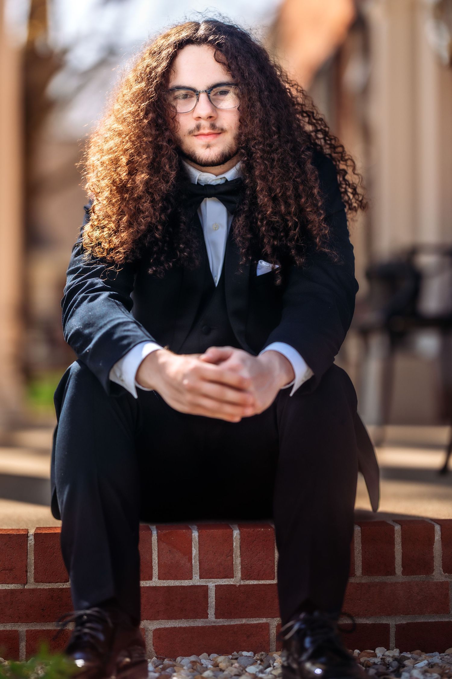 A person in formal attire with long curly hair and glasses sits on brick steps wearing a black suit and bowtie.