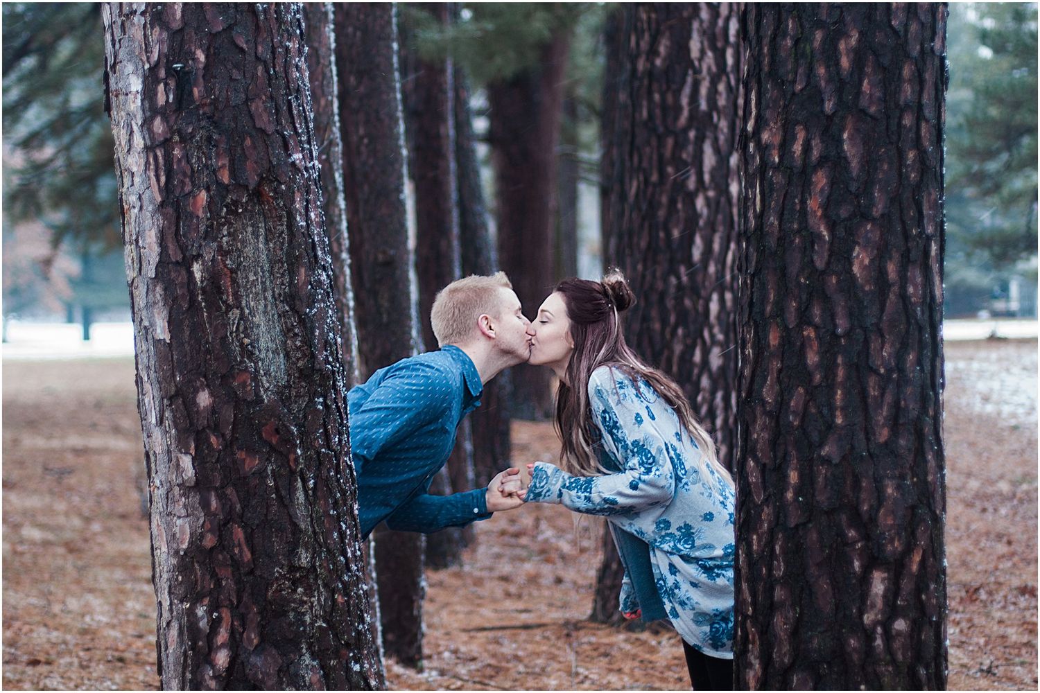 A playful kiss between a couple framed by tall pine trees in a winter forest setting.