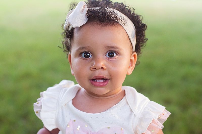 Adorable baby in pink and white dress smiling at camera outdoors.