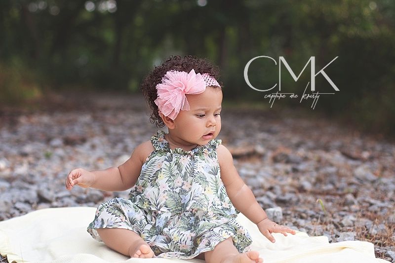 Baby wearing floral dress and pink bow sitting on white blanket with CMK watermark.