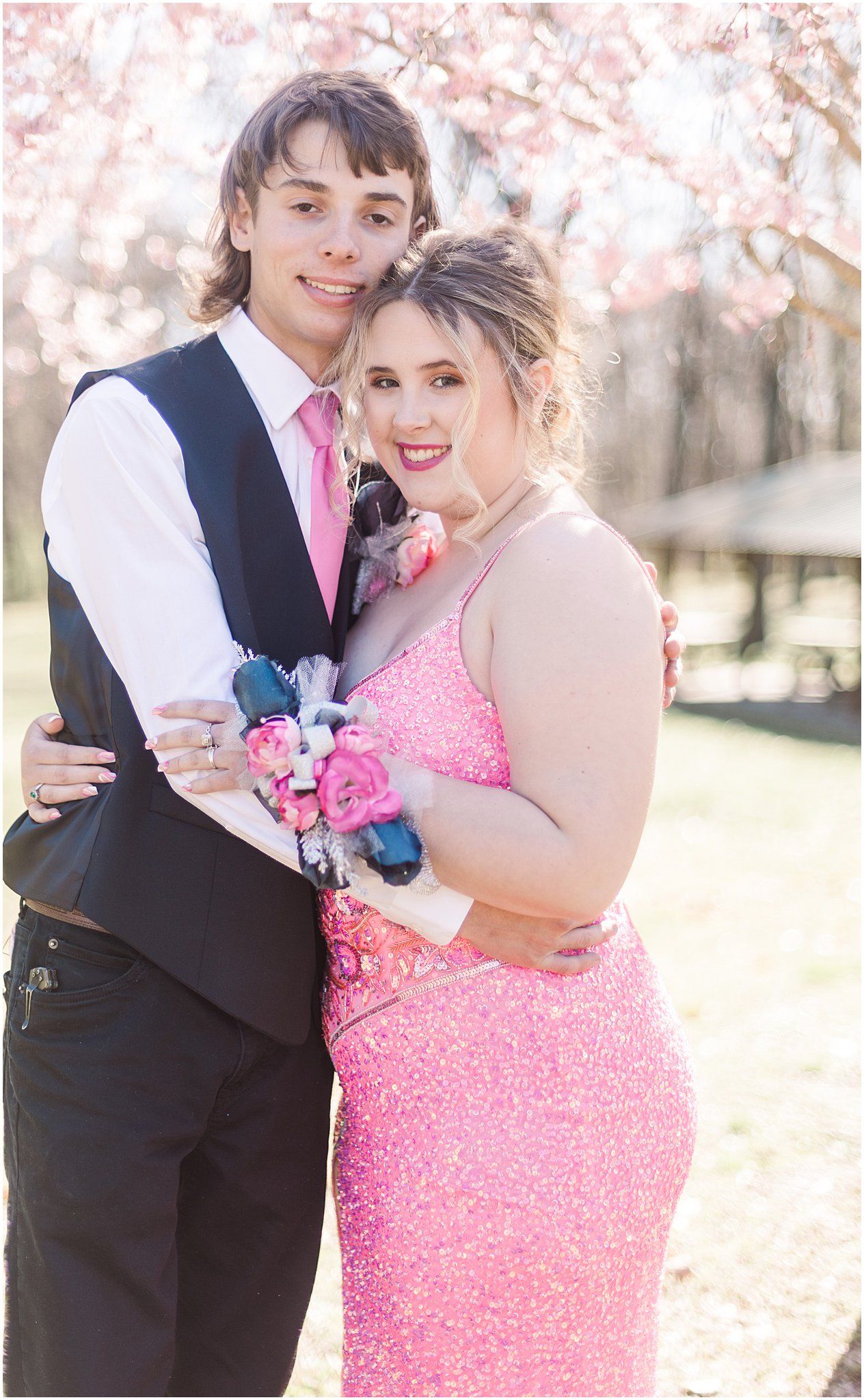 A couple in formal wear embraces under cherry blossoms, wearing navy vest and pink sequined gown.