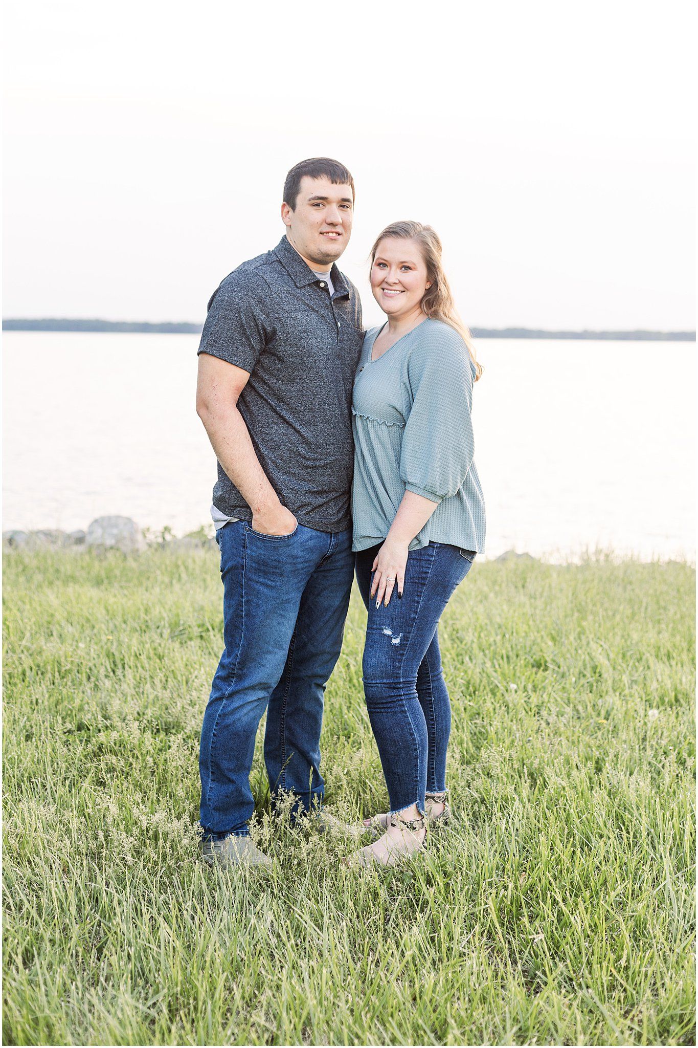 A couple stands together on grassy waterfront wearing coordinating casual outfits.