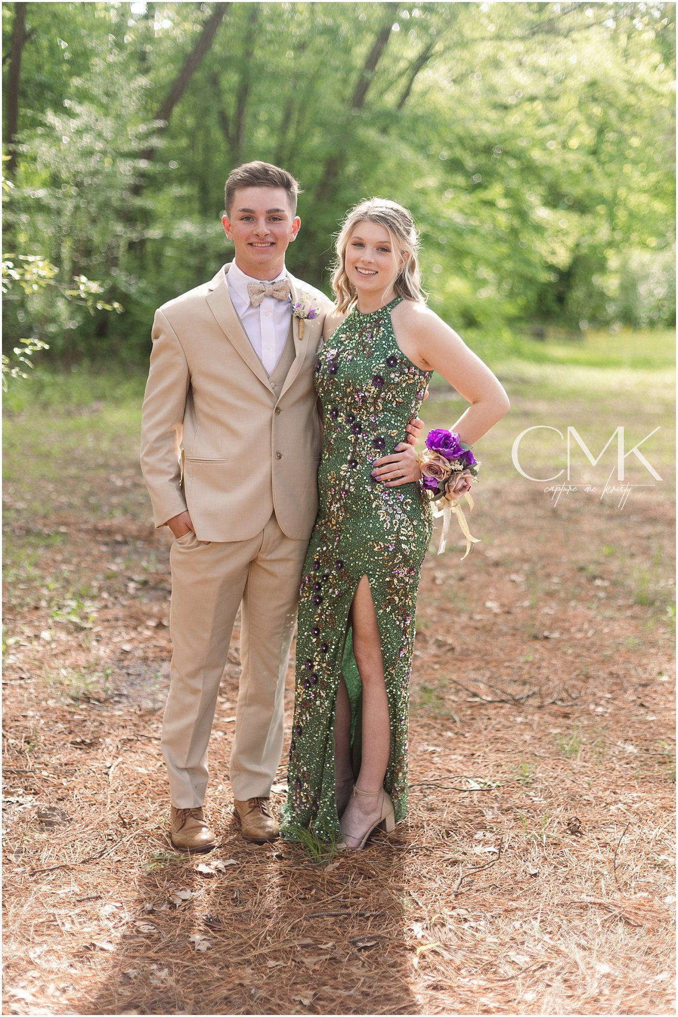 A formal couple poses in complementary tan suit and sparkling green gown with purple bouquet in woods.