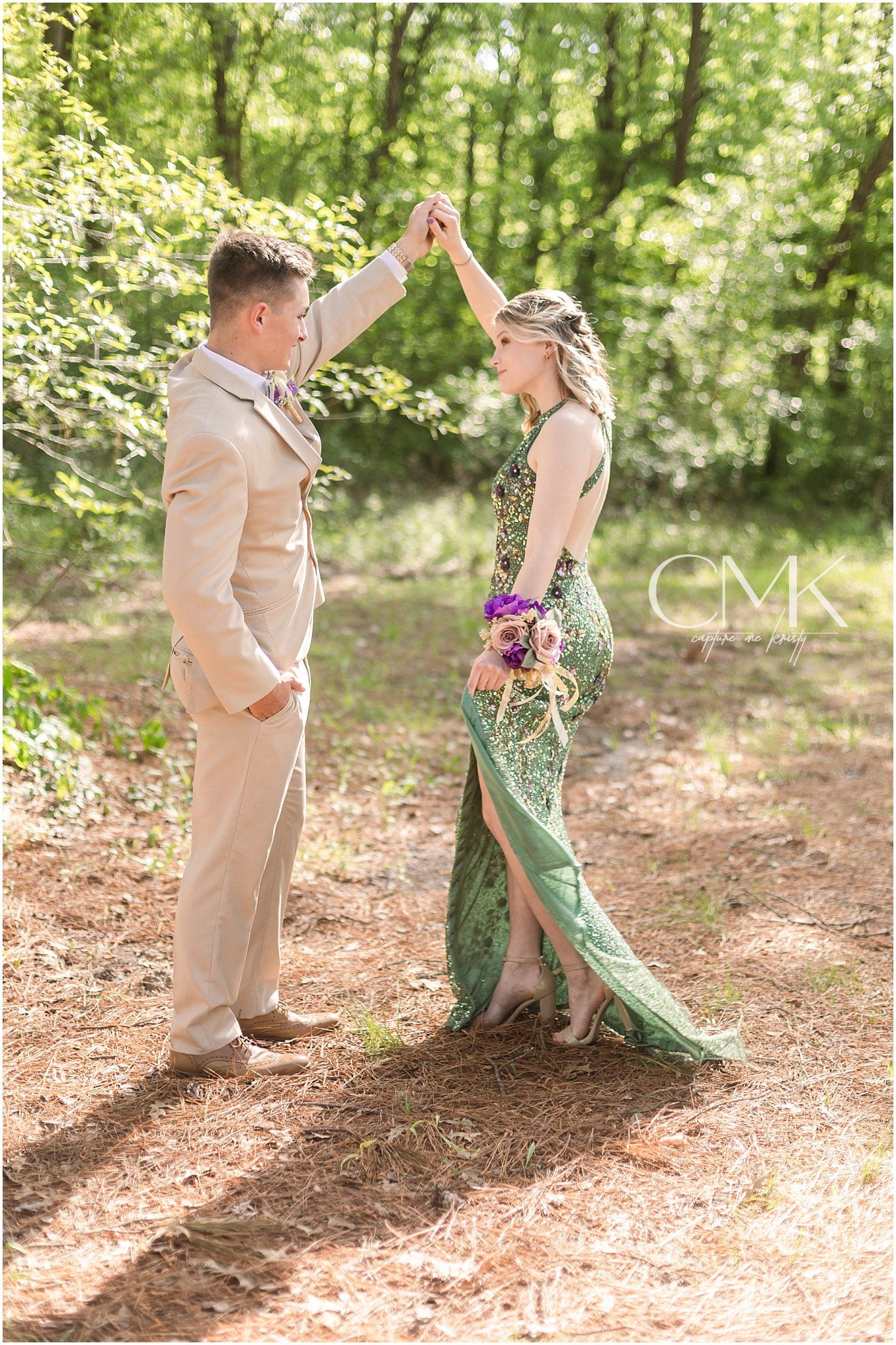 Couple dancing and holding hands in a forest wearing formal attire with flowing green gown.