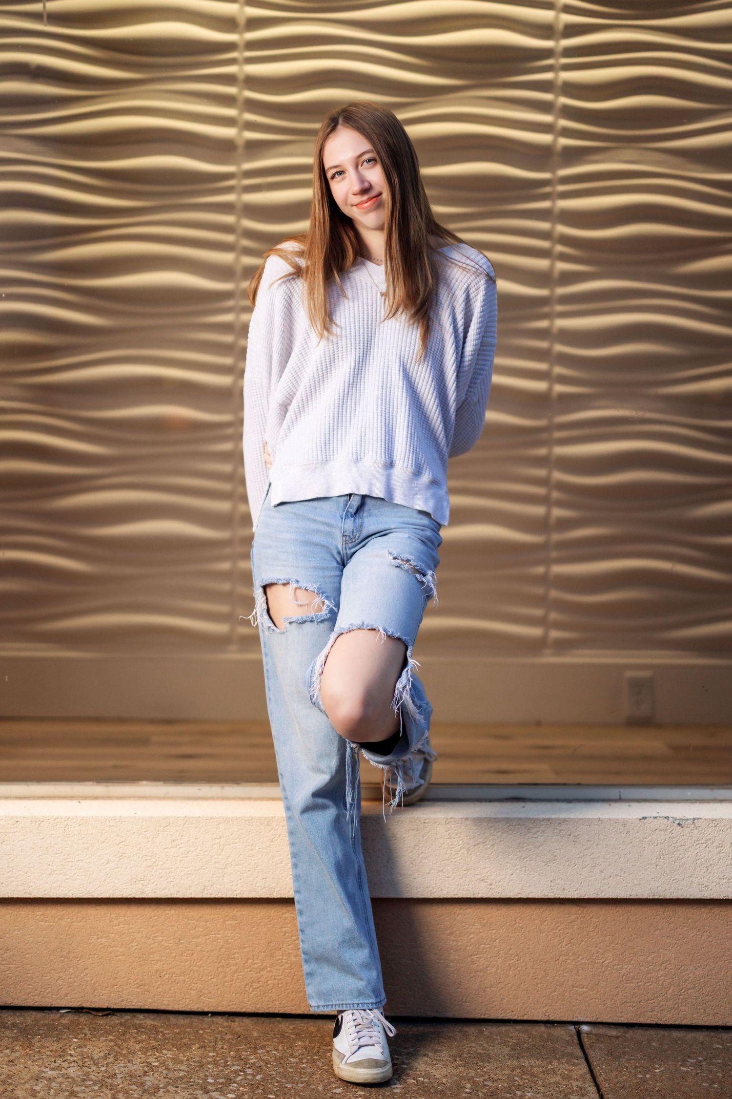 Person wearing ripped jeans and white sweater poses against textured gold wall.
