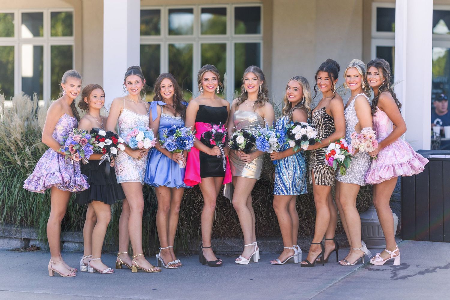 Group photo of nine friends in short party dresses holding colorful bouquets outside a building.