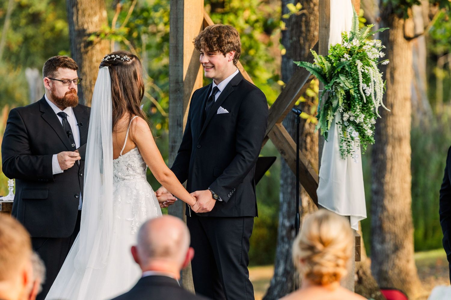 Wedding ceremony in progress under wooden pergola with floral decorations and gathered guests.