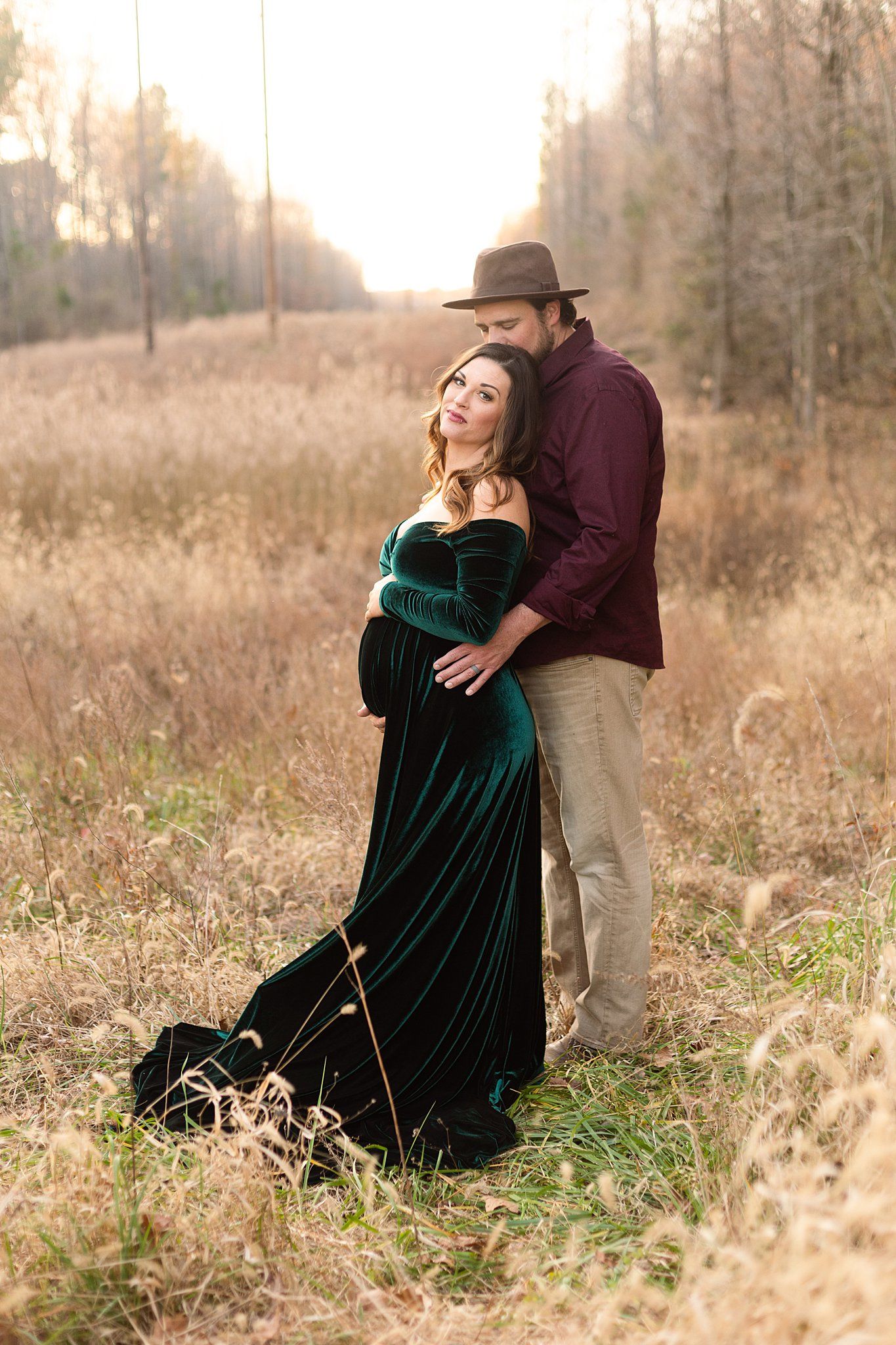 A couple embraces in a golden field at sunset with the woman wearing an elegant emerald velvet maternity gown.