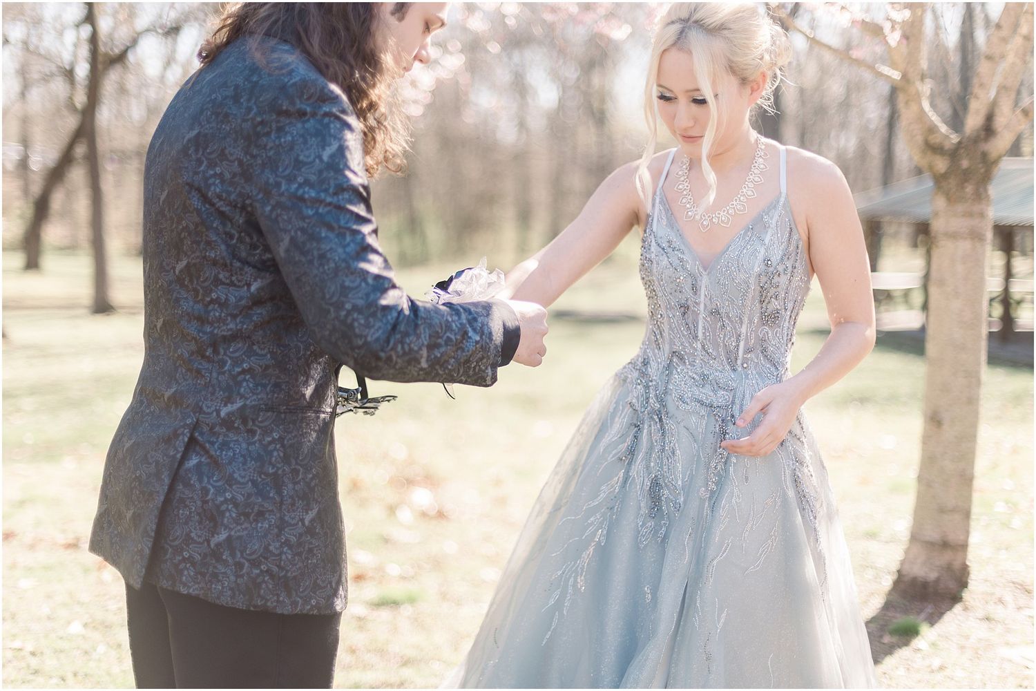 A couple in formal wear assist each other with clothing adjustments under blooming trees.