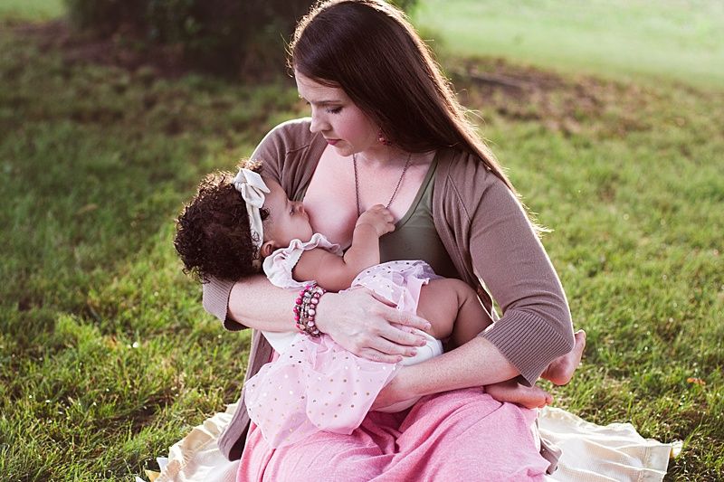 A tender breastfeeding moment captured outdoors on grass with soft natural lighting.