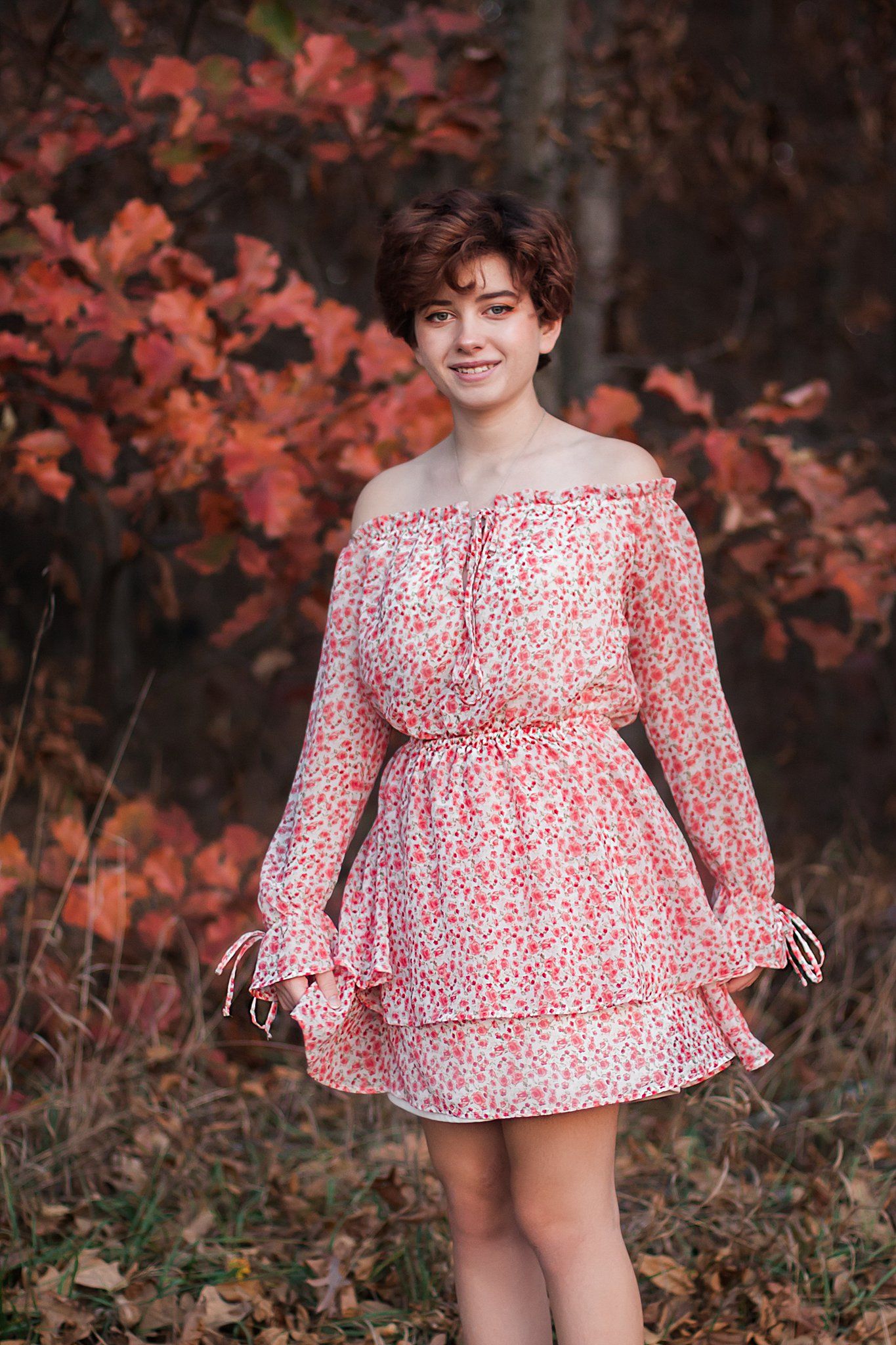 Person in floral off-shoulder dress poses against red autumn leaves.