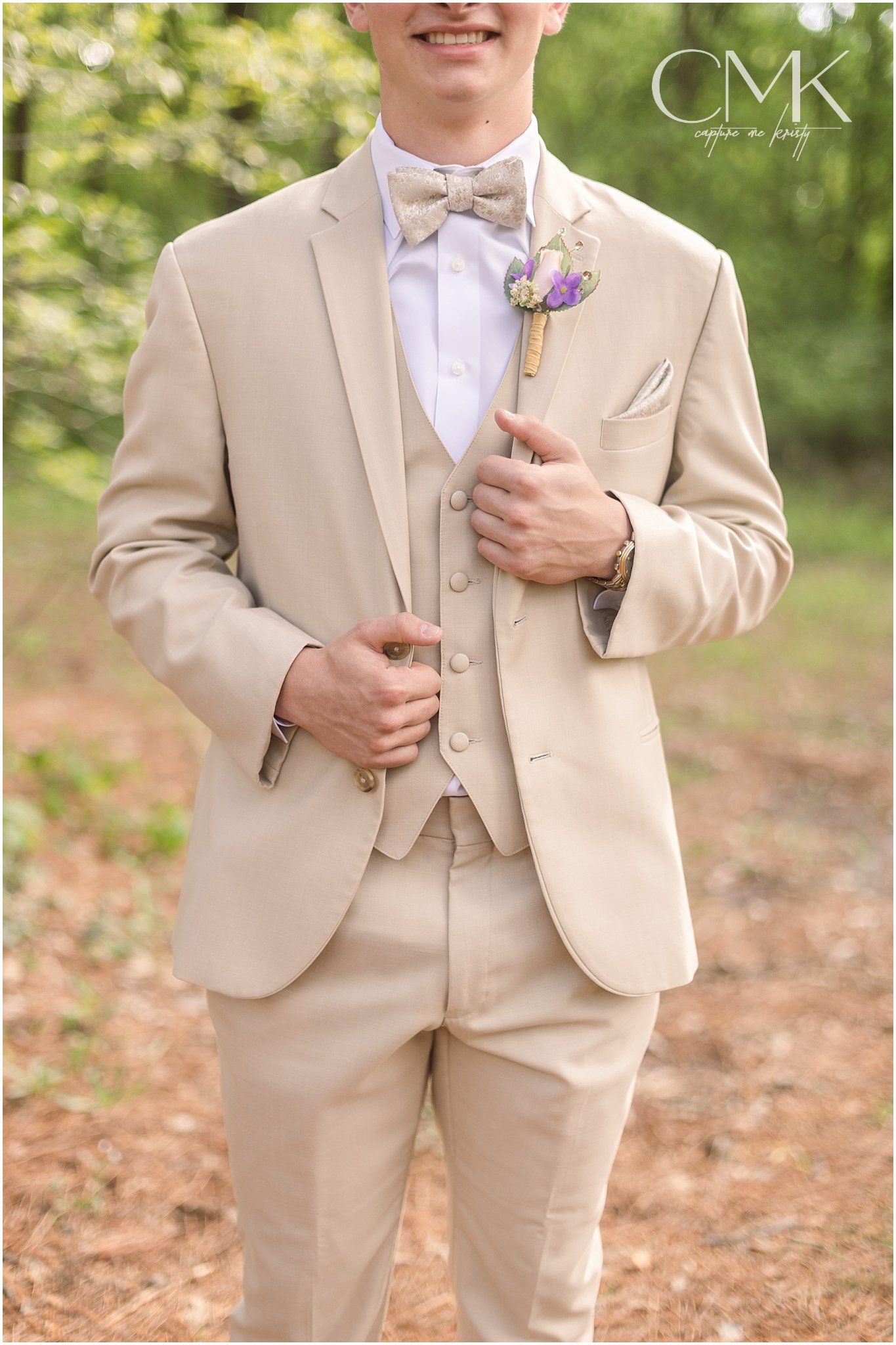 A three-piece tan suit with purple boutonniere and white bow tie against a green background.