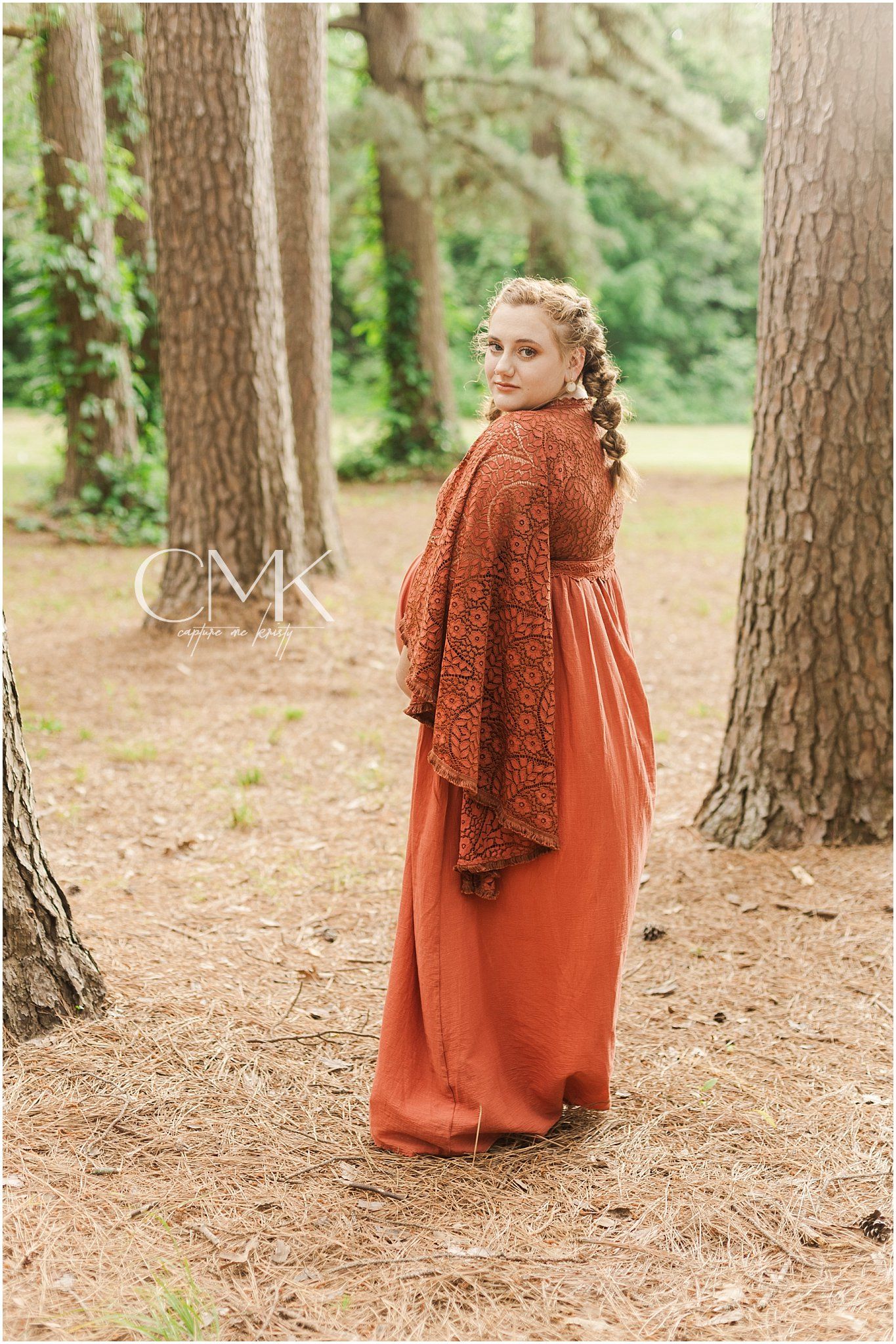 Full-length profile shot of an expectant mother in a rust-colored dress among pine trees.