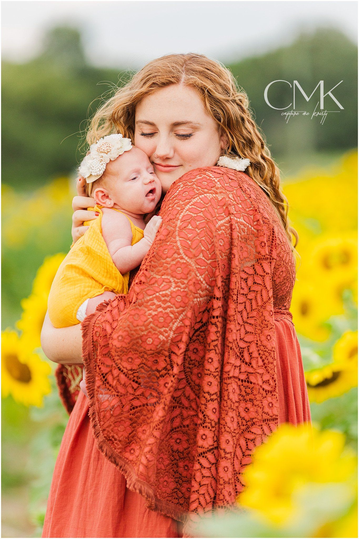 Mother in rustic lace dress embraces baby in yellow outfit among sunflower field.