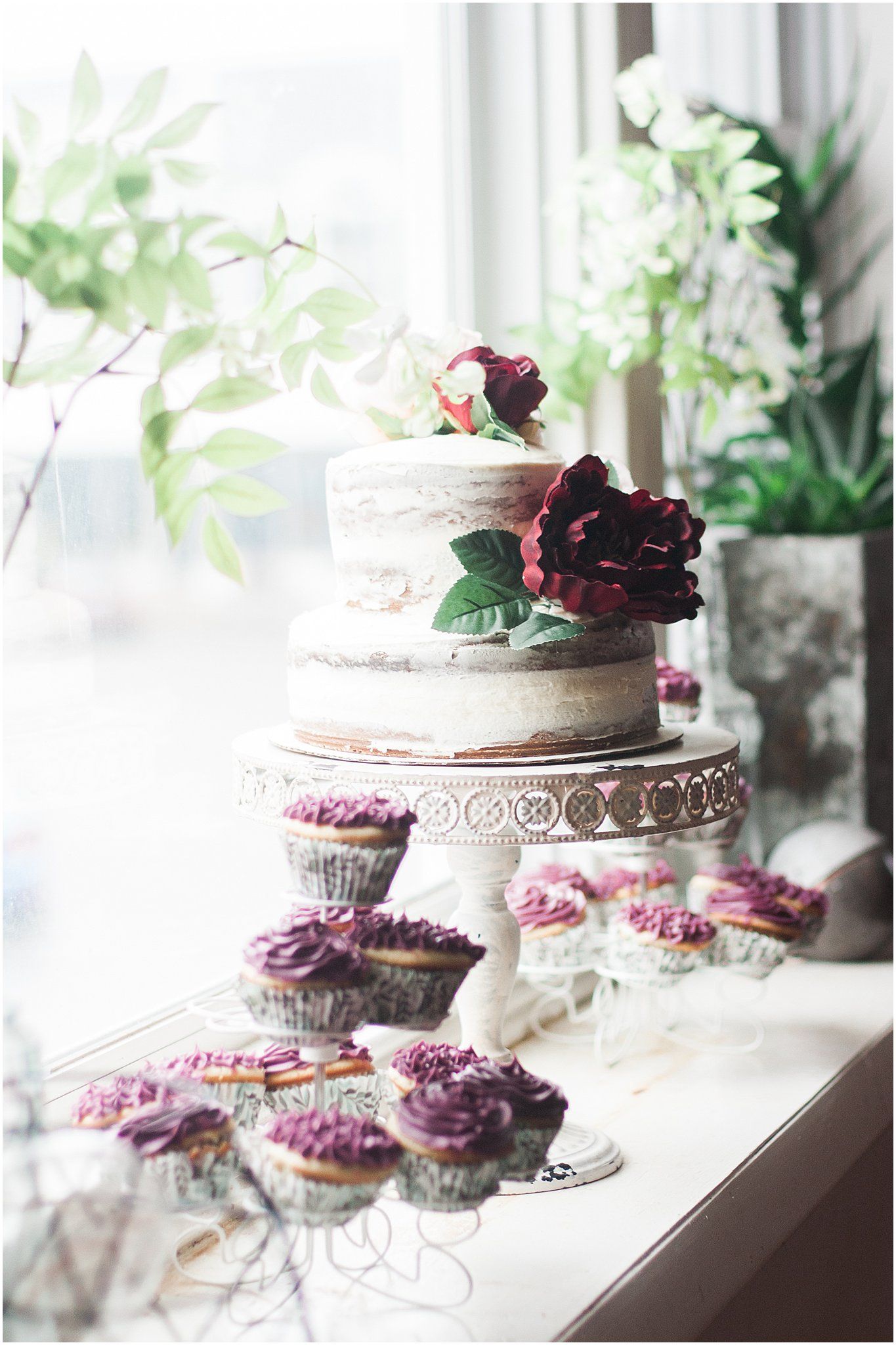 Romantic wedding cake setup with deep red roses and purple cupcakes by a bright window.