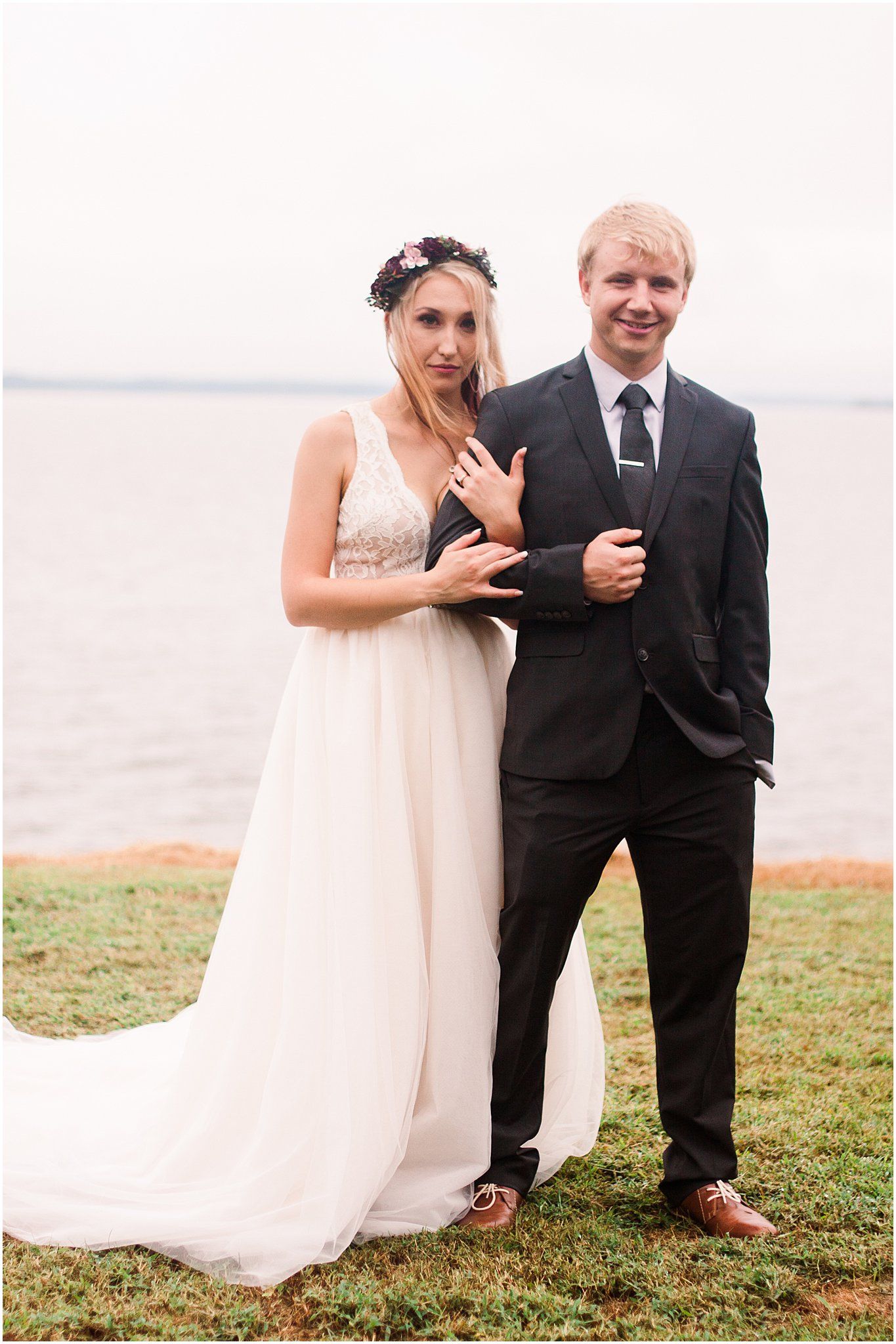 Happy couple poses together near a lake, with bride wearing a flowing dress and floral crown.