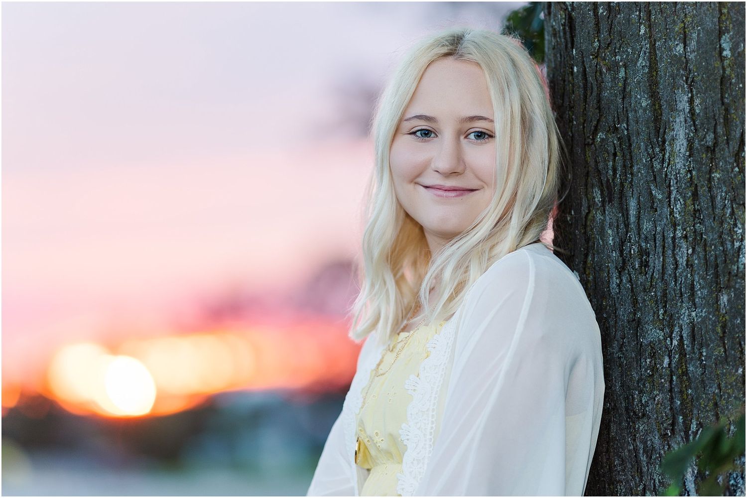 Senior girl leaning against a tree by a corn field with a pink and orange sunset in the background