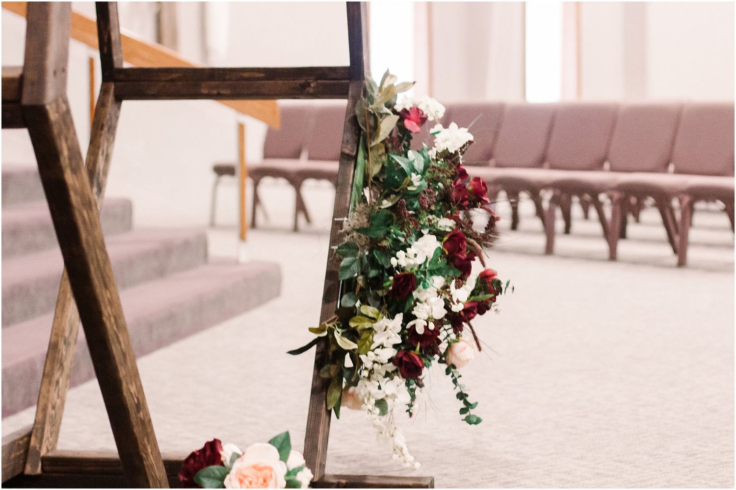 Rustic wooden ladder decorated with pink and burgundy floral arrangements in church setting.
