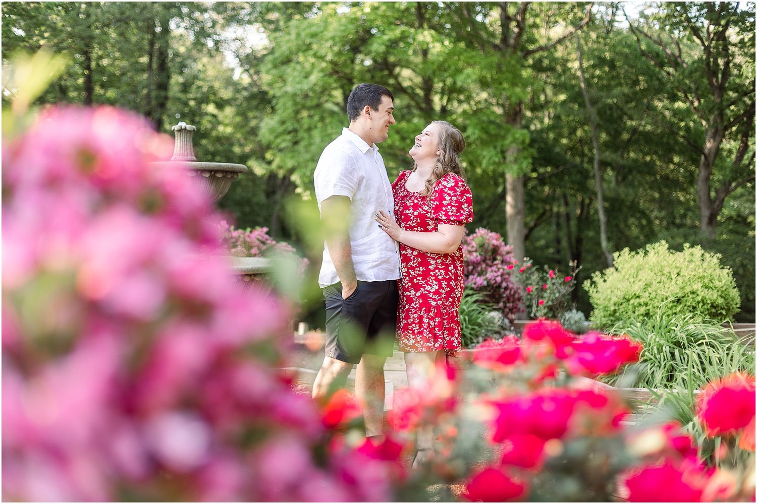 Romantic garden scene with couple among blooming pink flowers near decorative fountain.