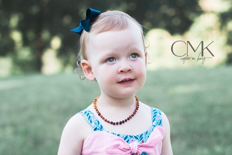 A toddler in pink and blue dress with bow and necklace smiles outdoors.