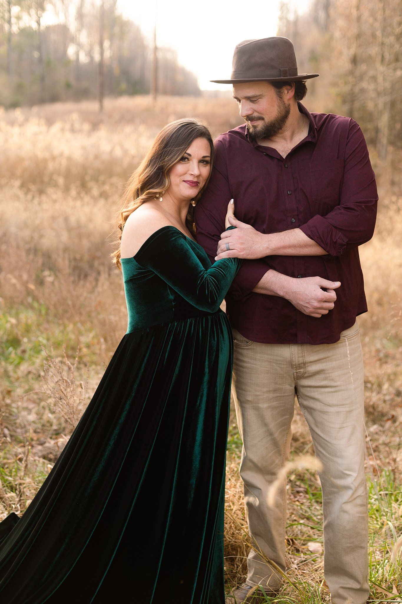 Maternity portrait with couple embracing in field wearing emerald gown and burgundy shirt.