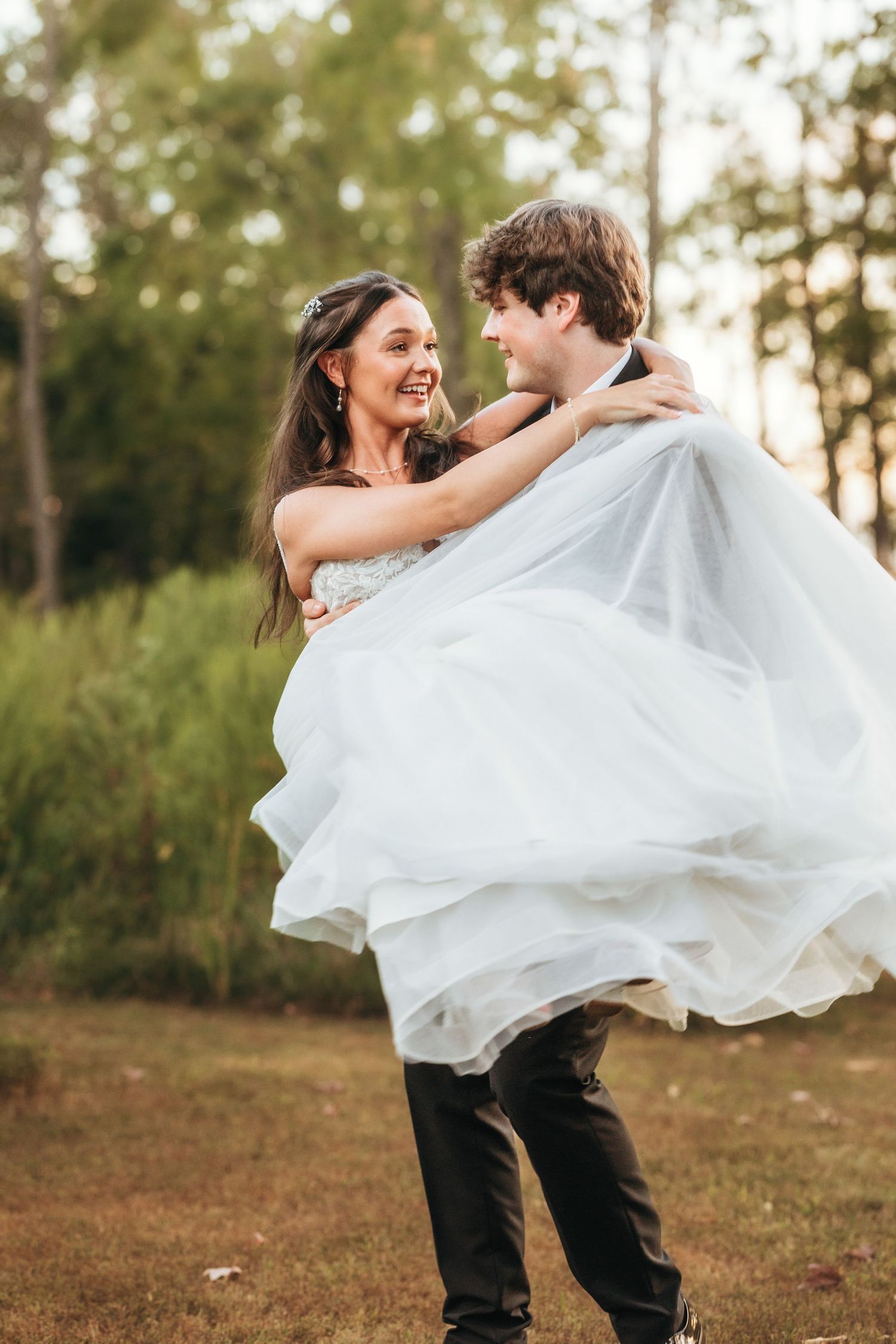 A joyful wedding moment with the groom carrying his bride in a flowing white dress against a natural backdrop.