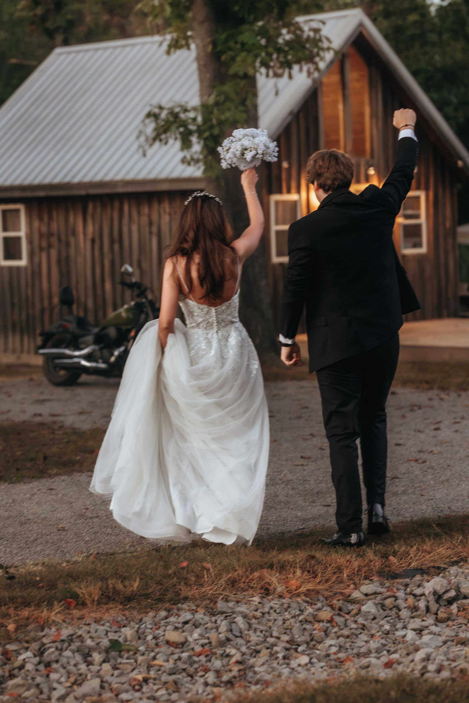 Newlyweds celebrate with raised arms while walking away from rustic wooden cabin.