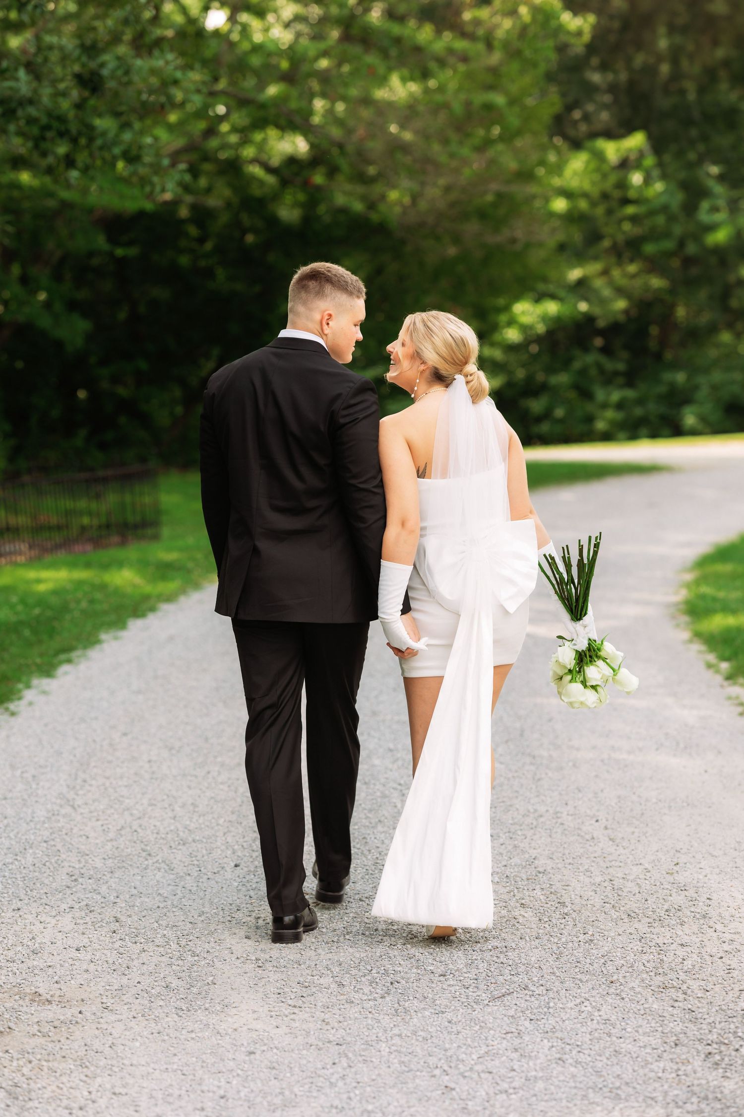 Newlyweds walk together on a gravel path surrounded by lush green gardens.