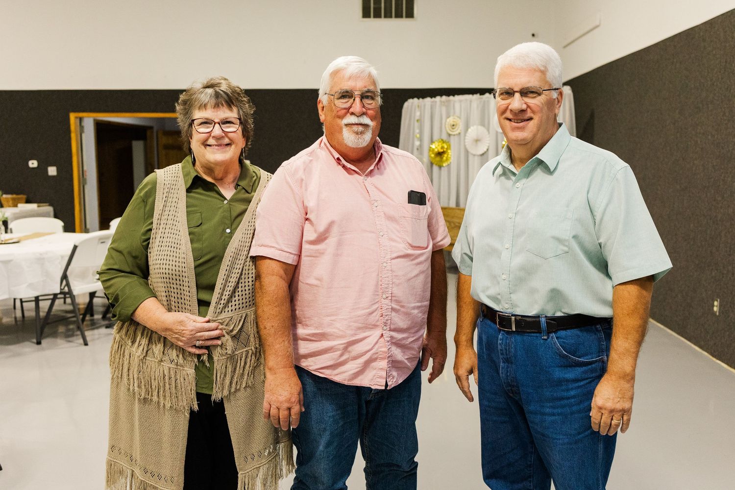 A group of senior adults stand together smiling in casual clothing at an indoor gathering.