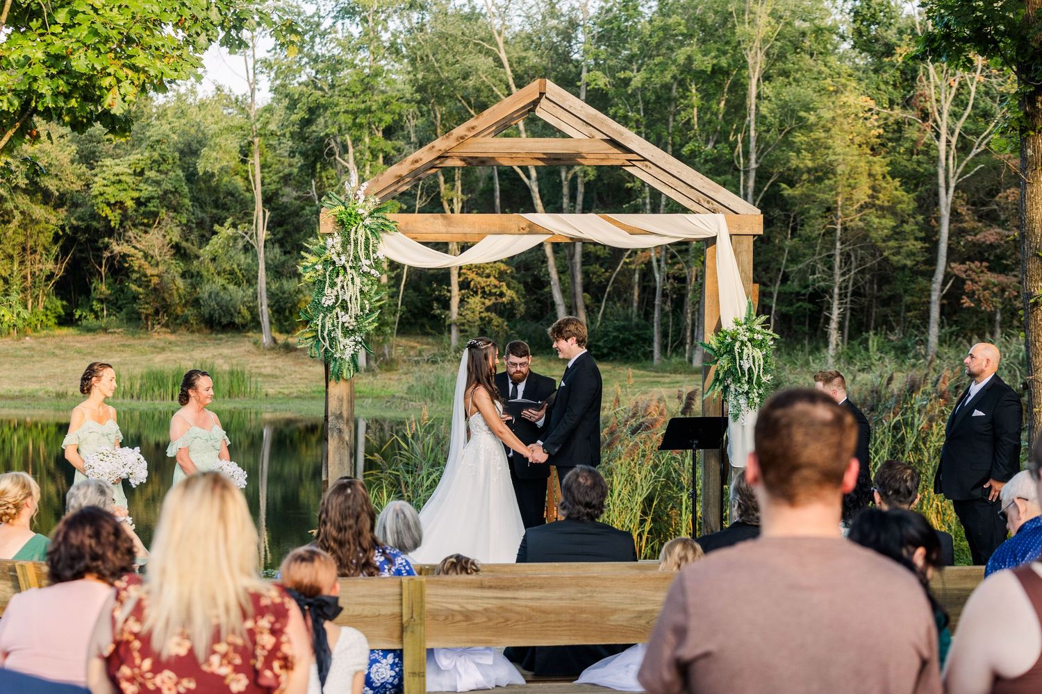 Outdoor wedding ceremony under a wooden arch with guests seated on white chairs.