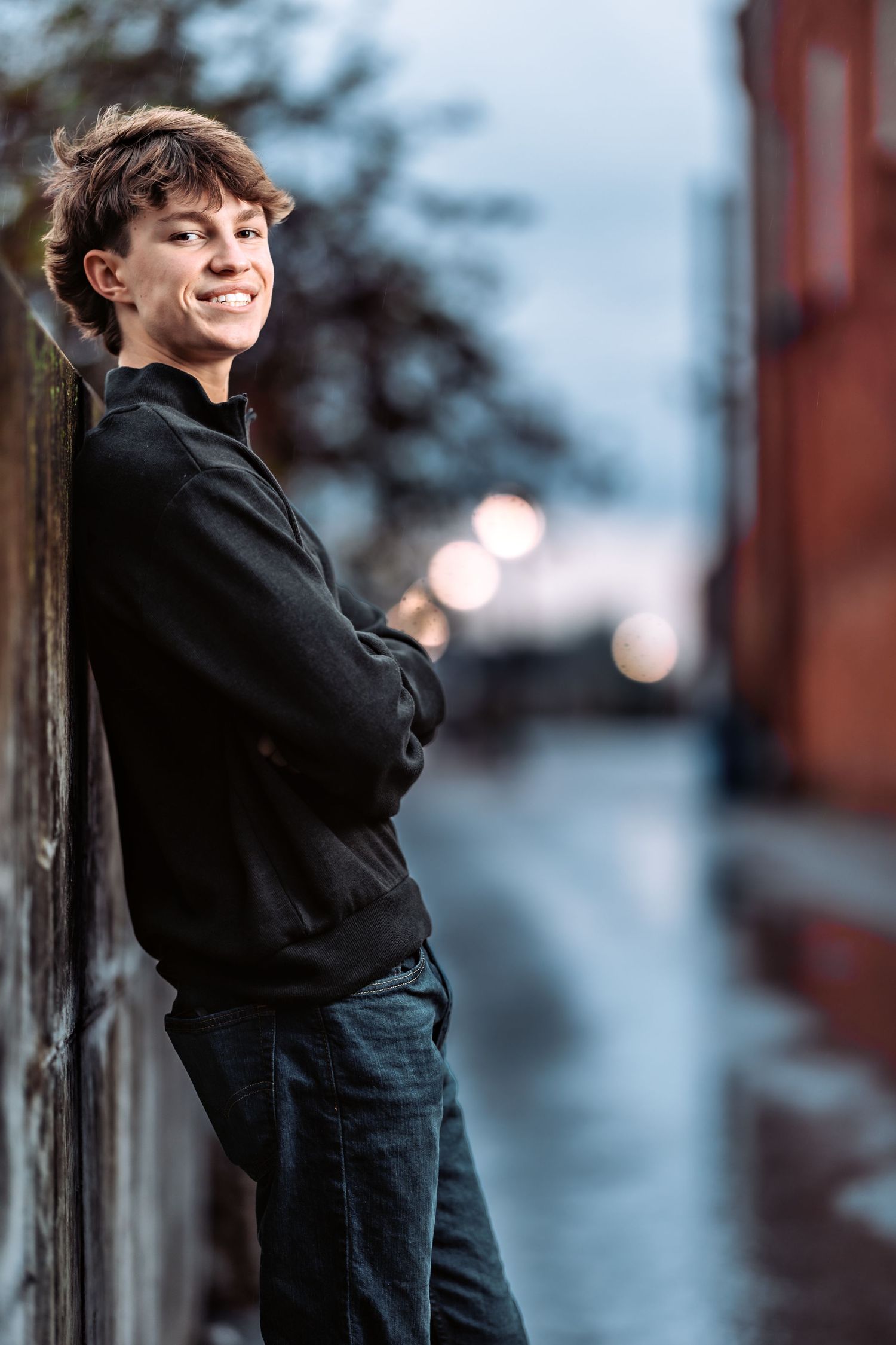 A person in a black coat and jeans leans against a wall in a moody blue-tinted urban setting.