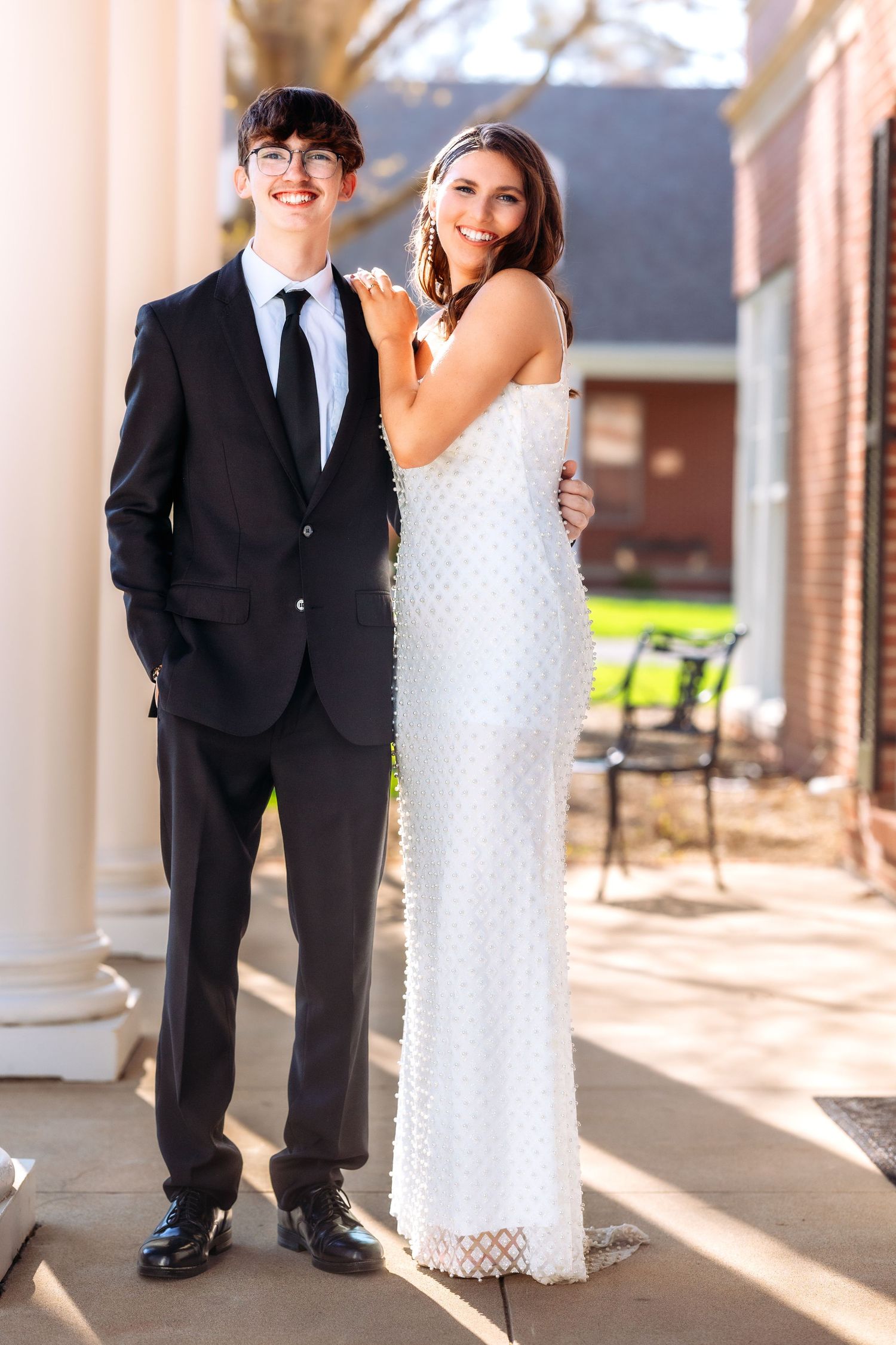 A couple in formal attire stands in a sunlit colonnade with white pillars, the groom in black and bride in white lace.
