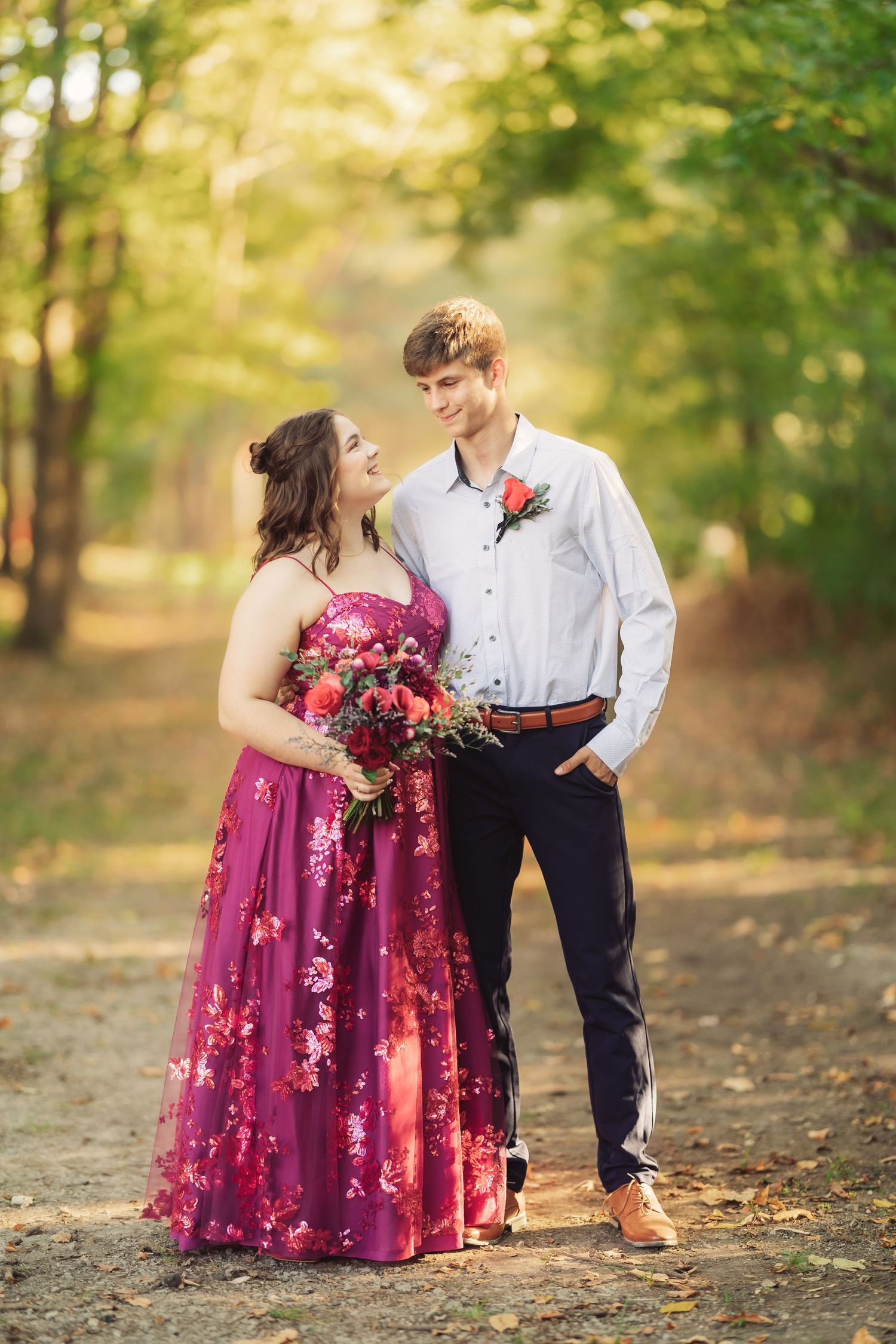 Couple posing together in formal attire with burgundy floral dress in an autumn forest setting.