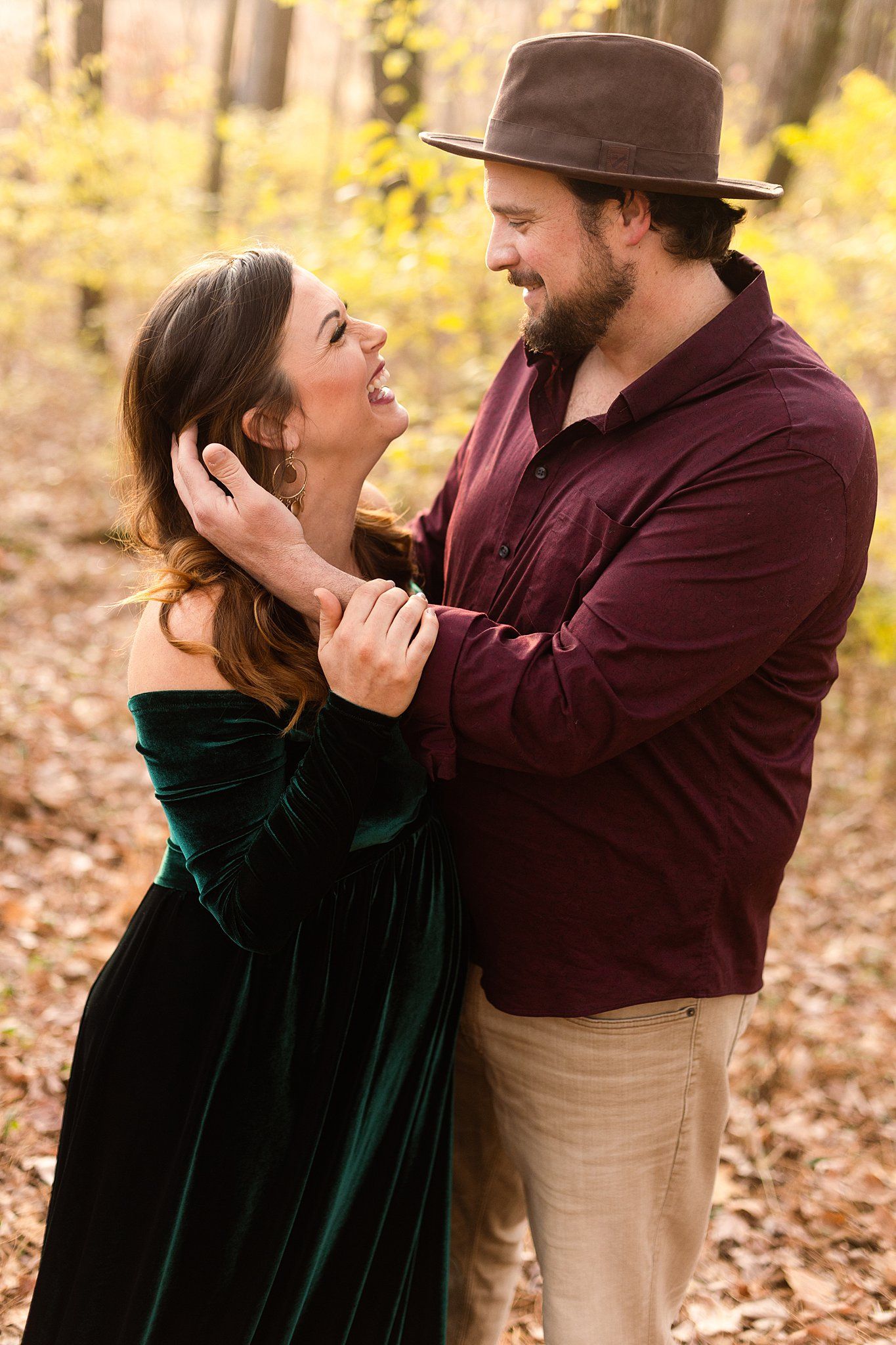 Joyful couple sharing a laugh in forest setting wearing burgundy shirt and green velvet dress.
