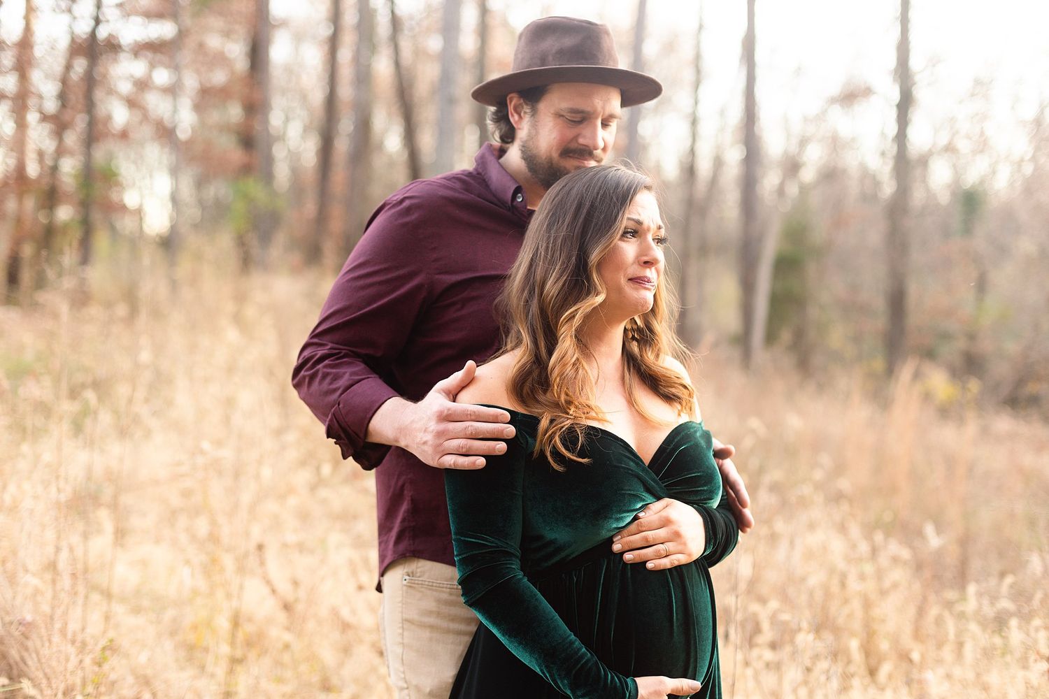 Romantic outdoor maternity photo with couple in burgundy shirt and emerald dress among fall foliage.