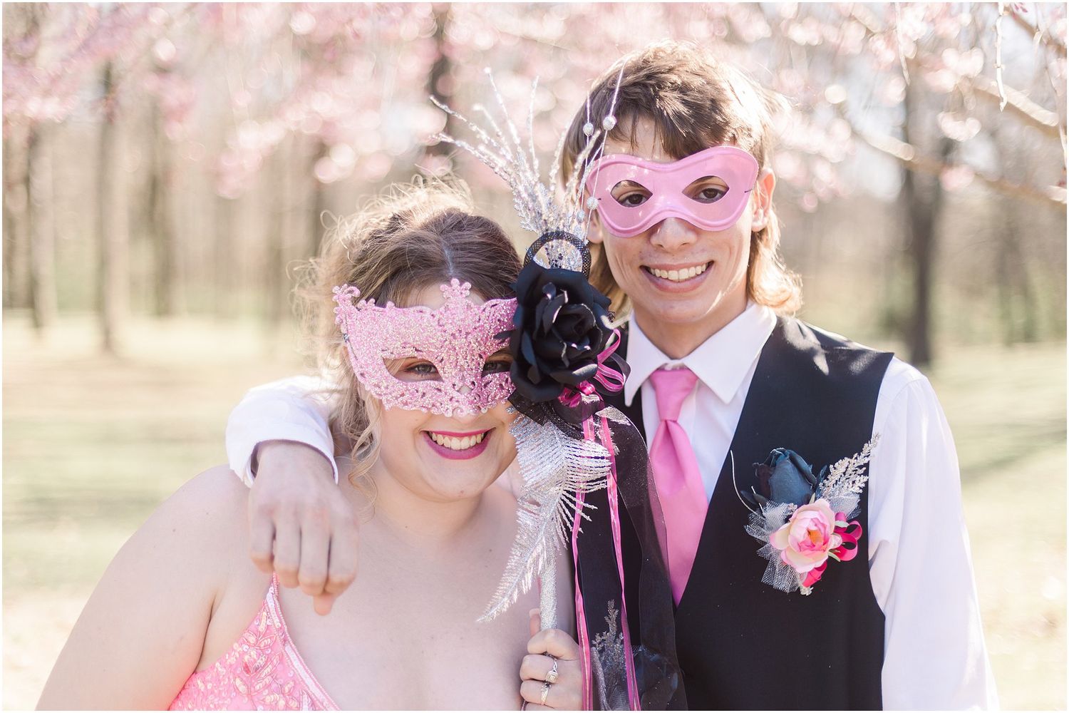 A couple wearing pink and black formal attire with matching masquerade masks pose together outdoors.