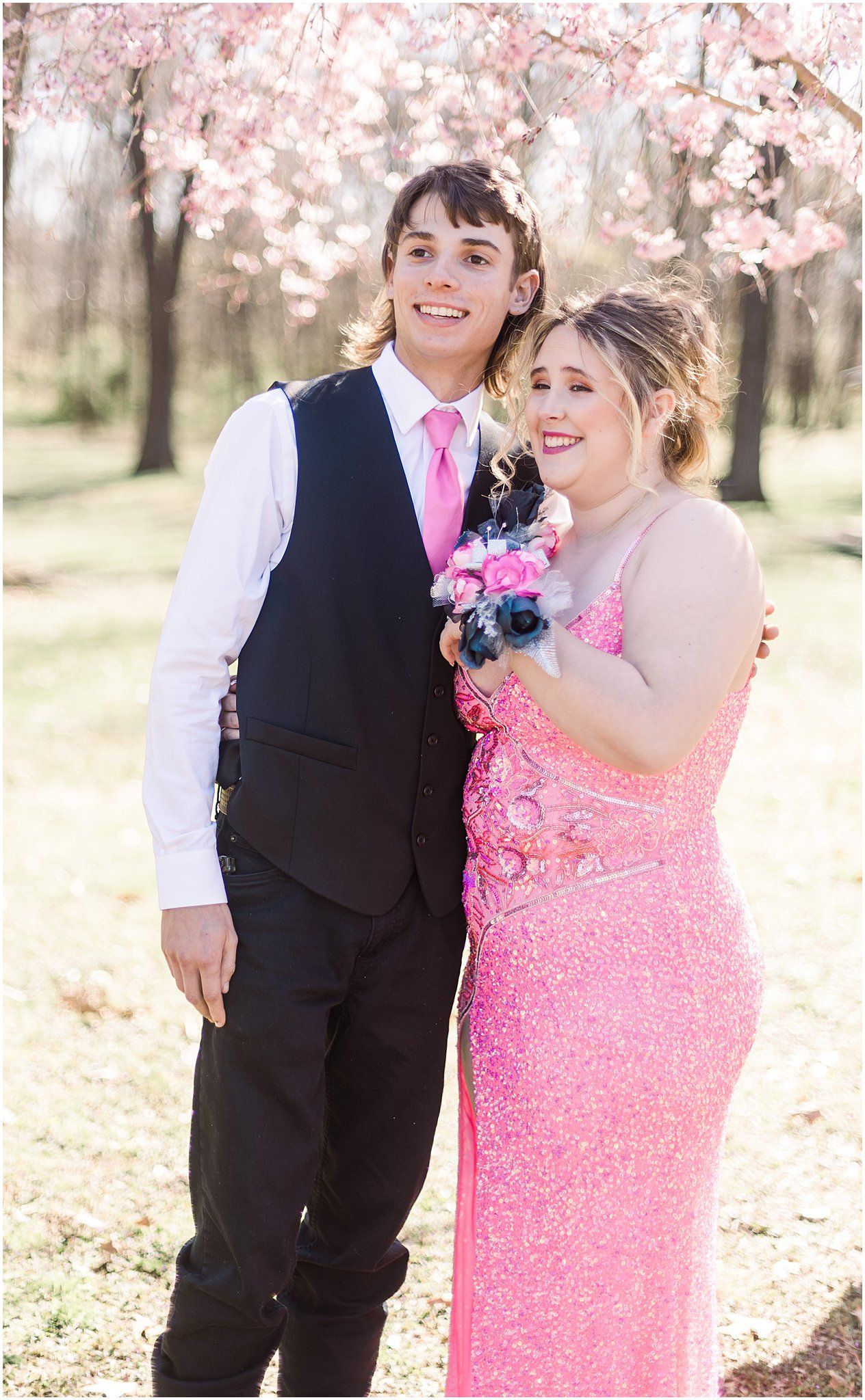 A couple smiles at the camera in formal wear with pink cherry blossoms in the background.