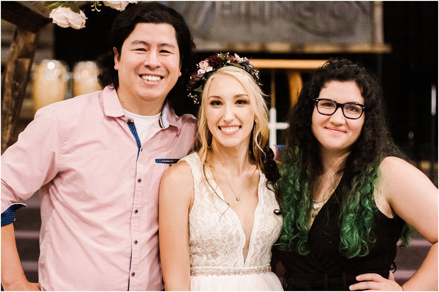 Three people smile at a rustic venue entrance, with the bride in a white dress and floral crown.