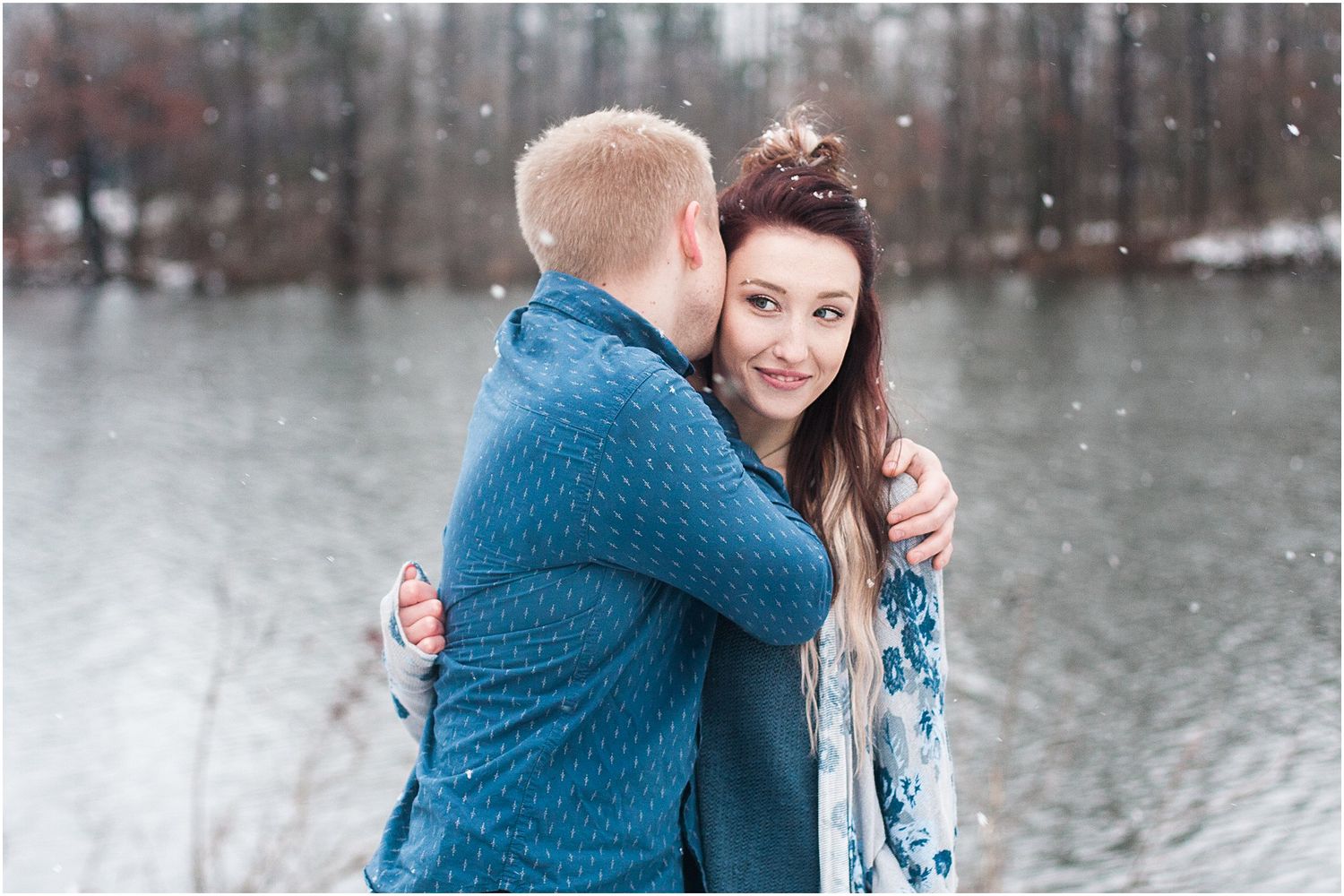 A tender embrace captured during a winter photoshoot with snow falling near a lake.