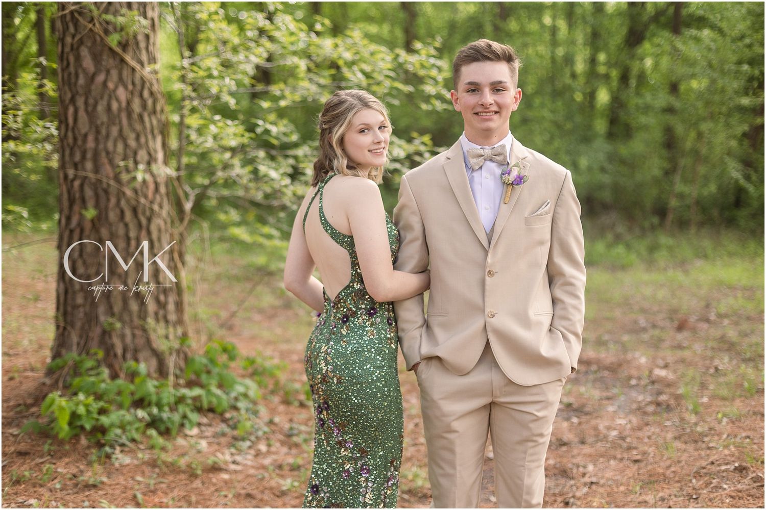 Forest prom photo of a couple in tan suit and sparkling green dress against natural backdrop.