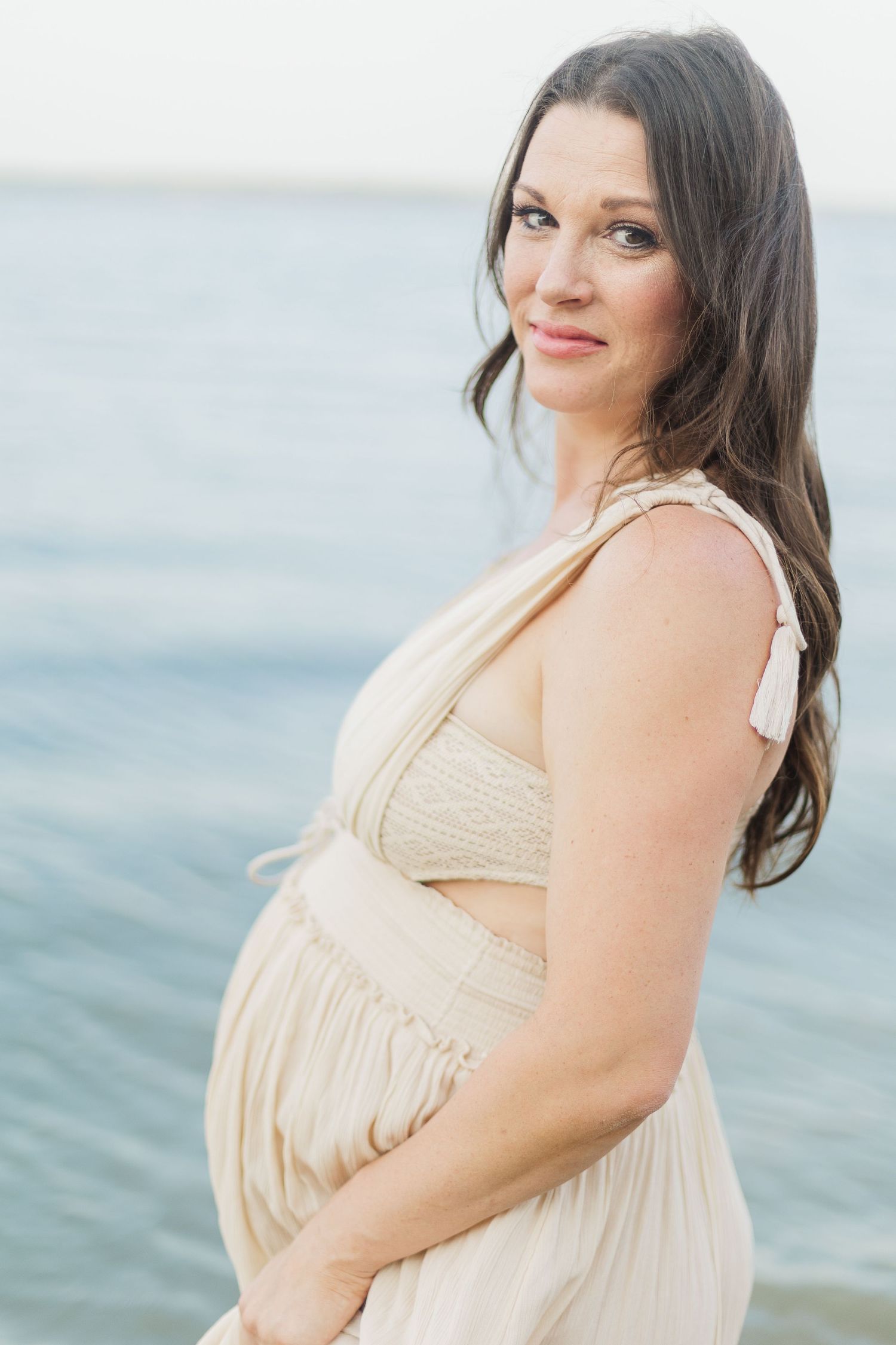 Woman in beige dress stands against ocean backdrop during golden hour.