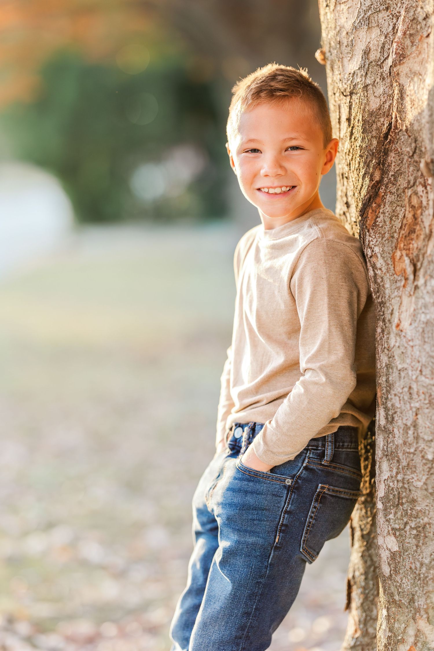 Young child in beige sweater and jeans poses playfully against a tree trunk.