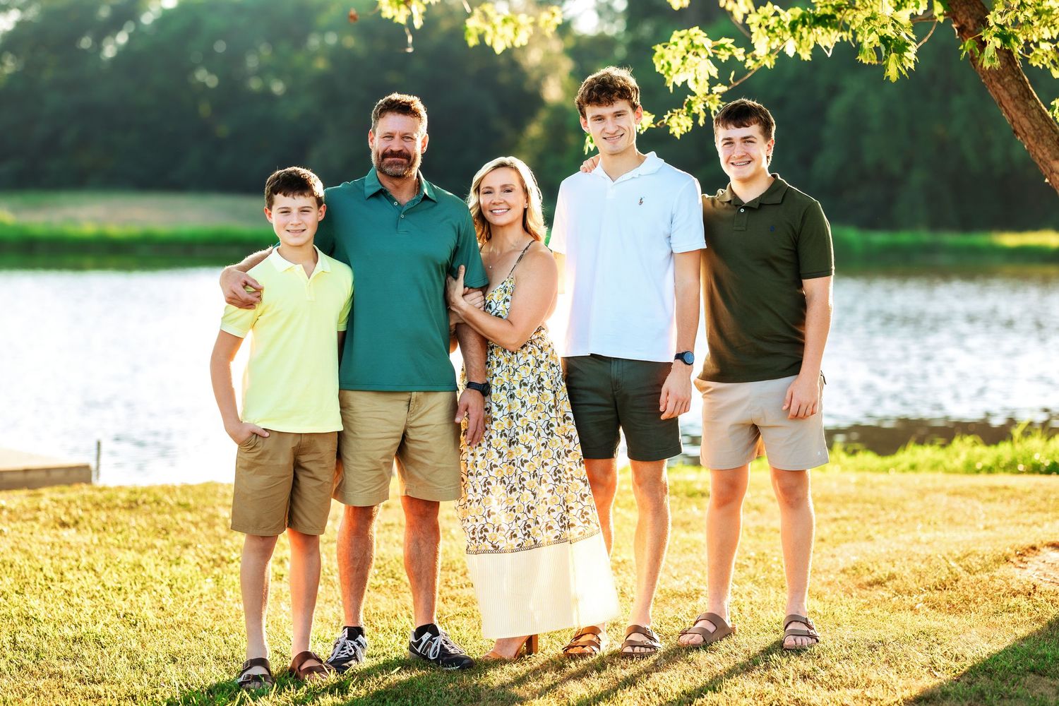 Family group stands together on grassy shore with lake and sunset in background.