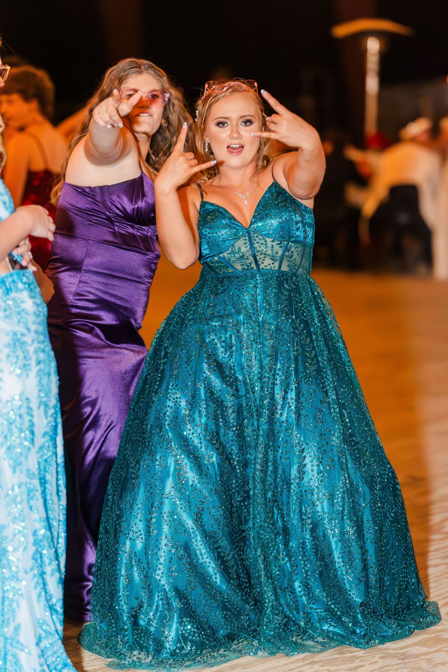 People in sparkling teal and purple formal gowns dance on a dance floor at a celebration.