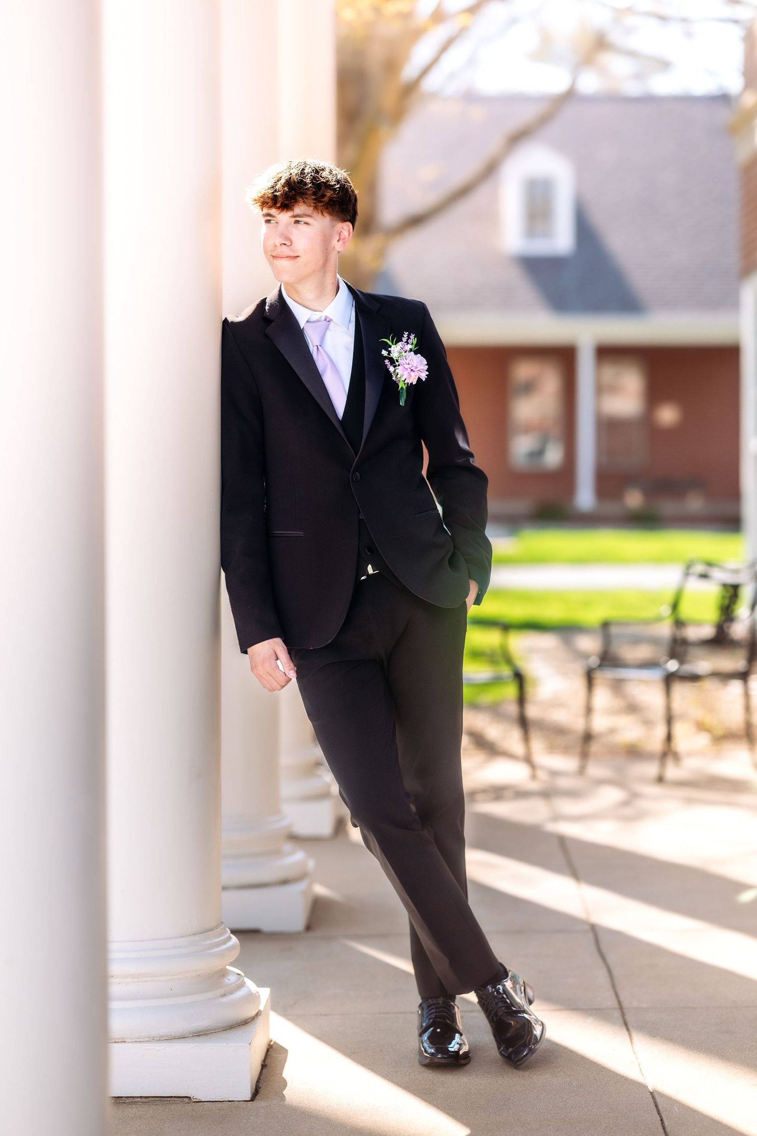 A person in a formal black tuxedo and pink boutonniere stands between white columns at an outdoor venue.