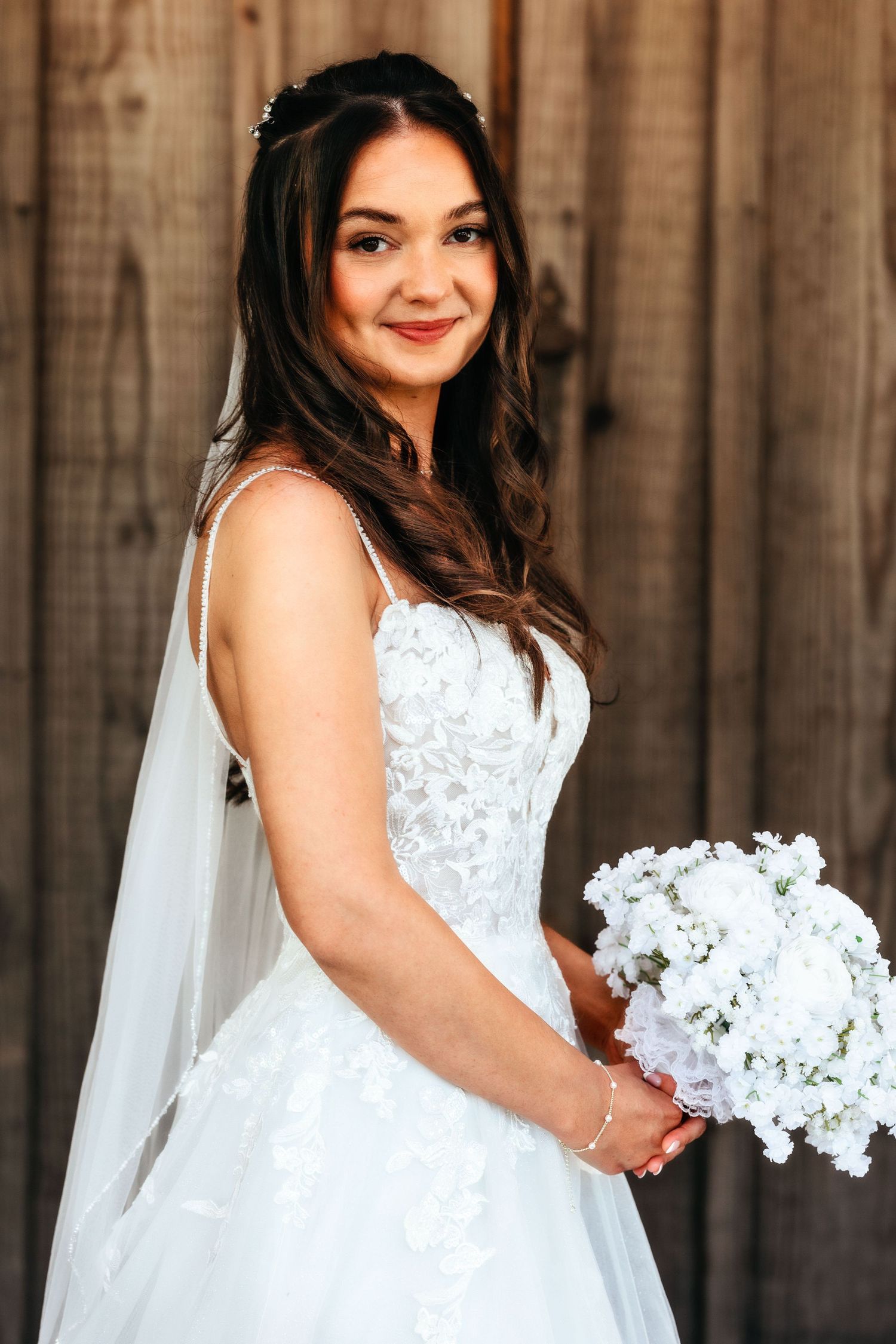 Bride in white lace wedding dress holds white floral bouquet against wooden backdrop.