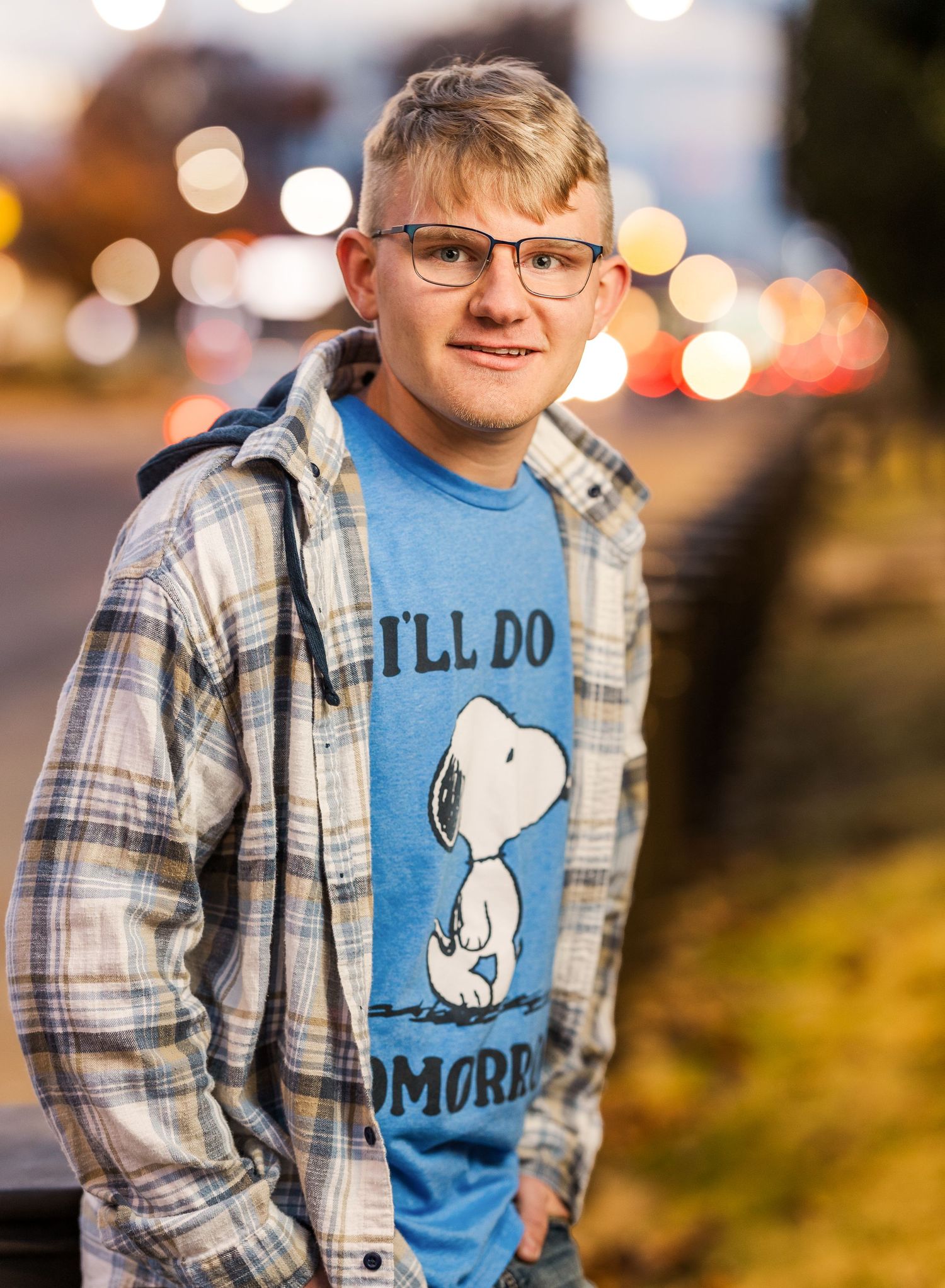 Young person wearing a plaid shirt and blue Snoopy t-shirt poses outdoors with bokeh lights in background.