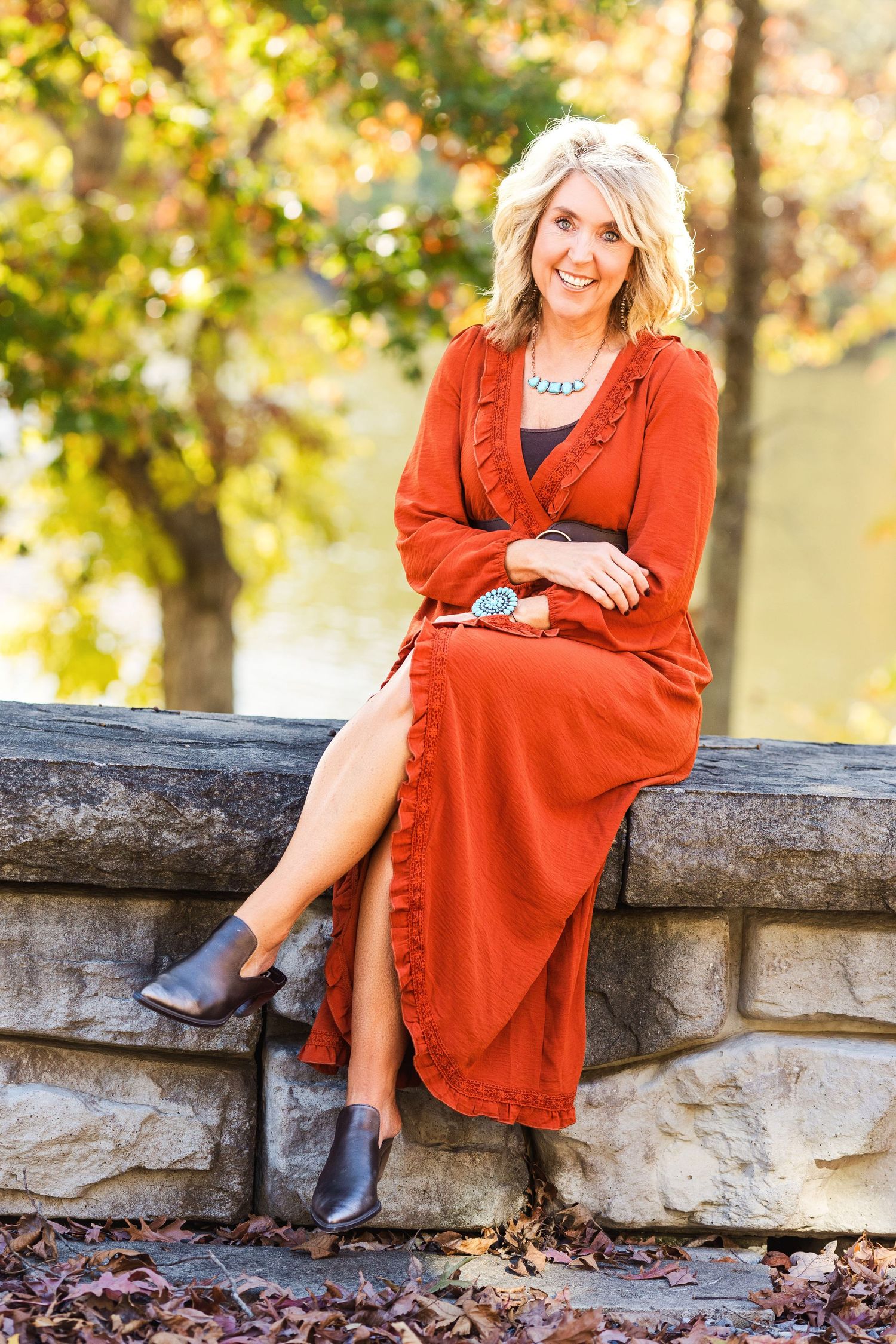 A portrait of someone wearing a stylish rust-colored dress while sitting on a stone wall in autumn.